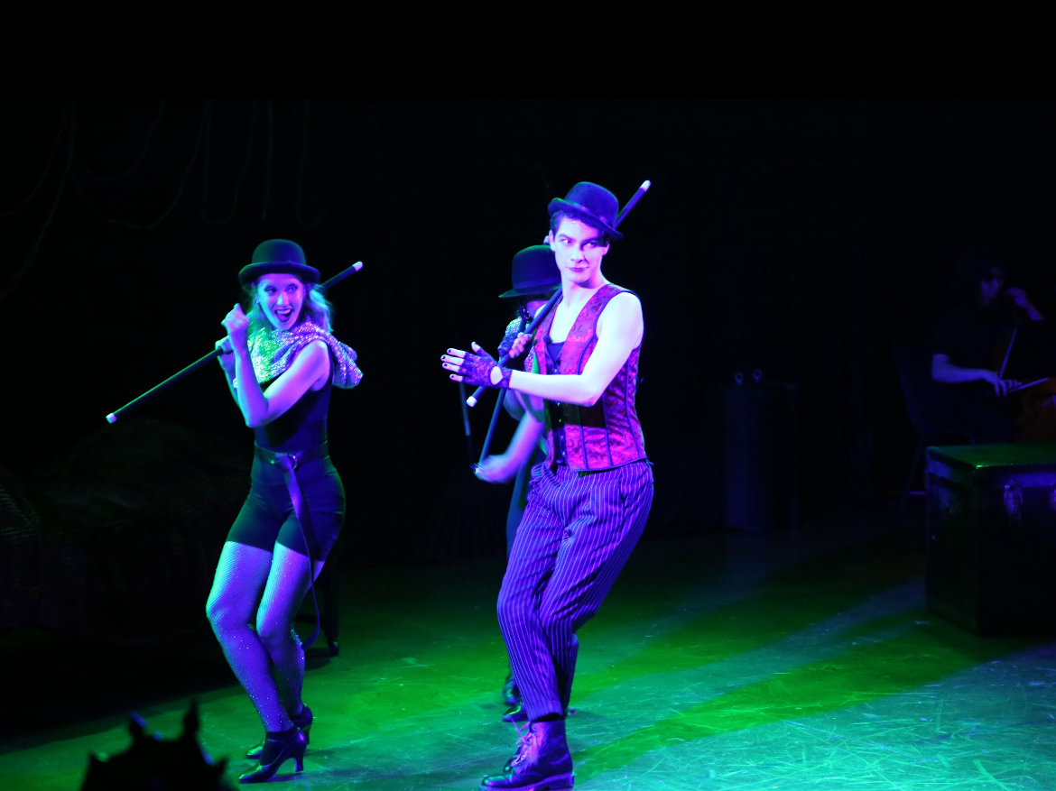 Performer dressed as a circus or cabaret artist in a colorful costume, wearing a top hat, gloves, and striped pants, holding a baton, on stage with a dark background and green stage lighting.