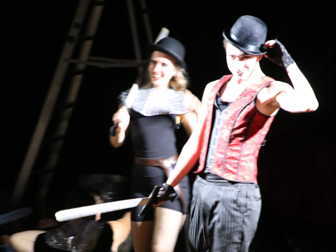 Two performers in vintage-style costumes, one with a top hat and the other with a bowler hat and a prop cane, on stage against a dark background.