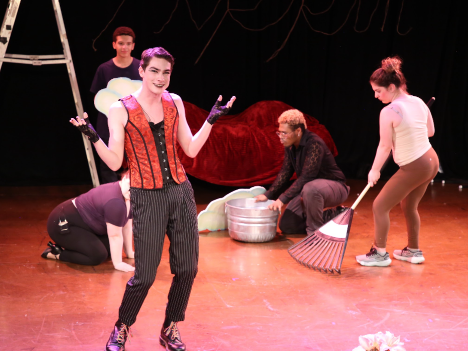 Group of people preparing a theatrical performance on stage, with one person smiling and gesturing to the camera and others tending to props in the background.