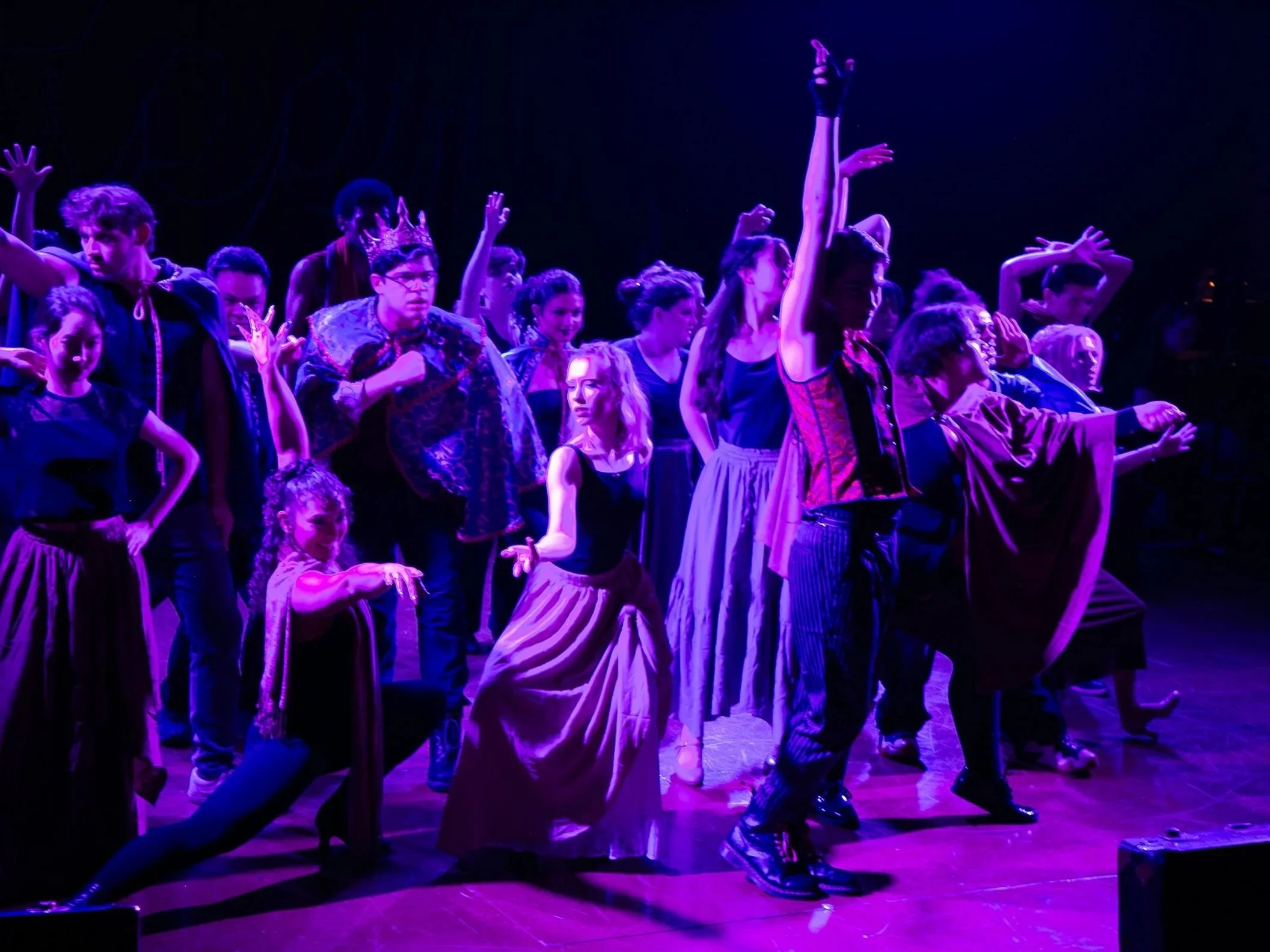 A group of people, both children and adults, performing a dance on stage under purple lighting. Some are raising their hands and displaying expressive movements, dressed in casual and theatrical costumes.