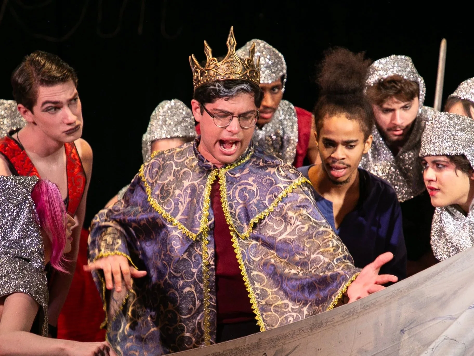 A group of actors in colorful costumes, with one wearing a crown and crown-shaped headpieces, gathered around a large book or script, on a stage with a black background, preparing for a theatrical performance.