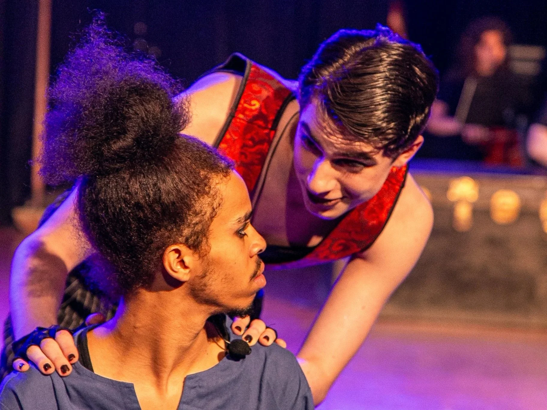 A performer with short dark hair in a red and black costume leaning over a seated man with a beard and long dark hair, in a setting with purple lighting.