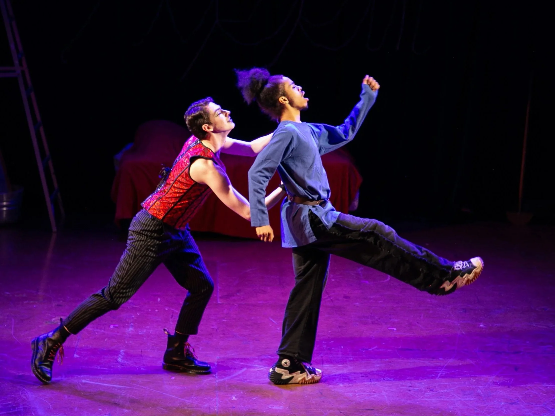 Two dancers are performing on stage, one behind the other, with one leg extended outward. They are dressed in colorful, casual clothing, with a dark background and stage lighting highlighting them.