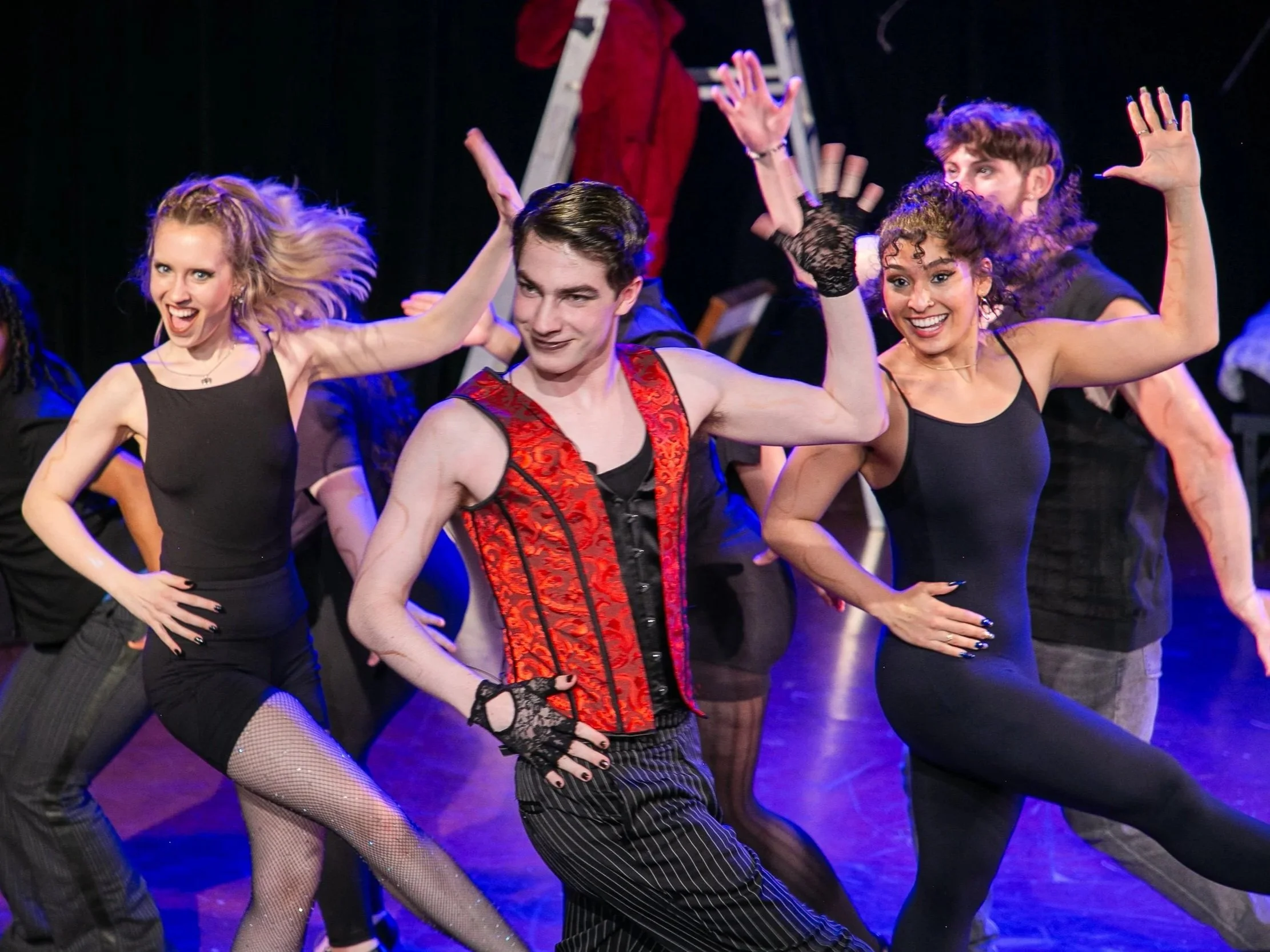 A group of performers on stage dancing with expressive poses, wearing costumes including fishnet stockings, gloves, and striped pants, with a dark background and stage lighting.