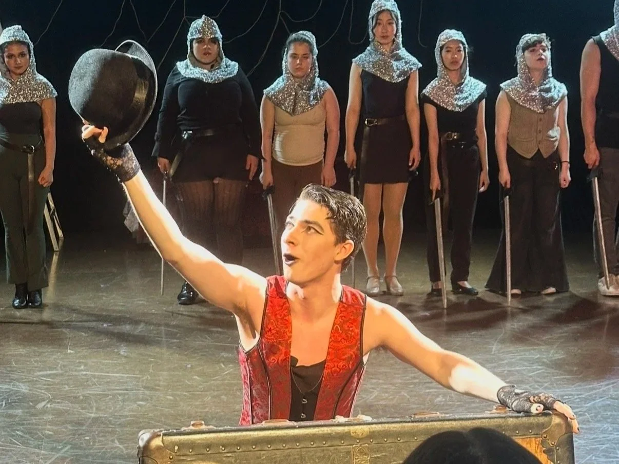 A male performer with dark hair, wearing a red and black costume, holding a black top hat in one hand and a cane in the other, performing on stage with a group of women dressed in black and silver costumes with silver head coverings in the background