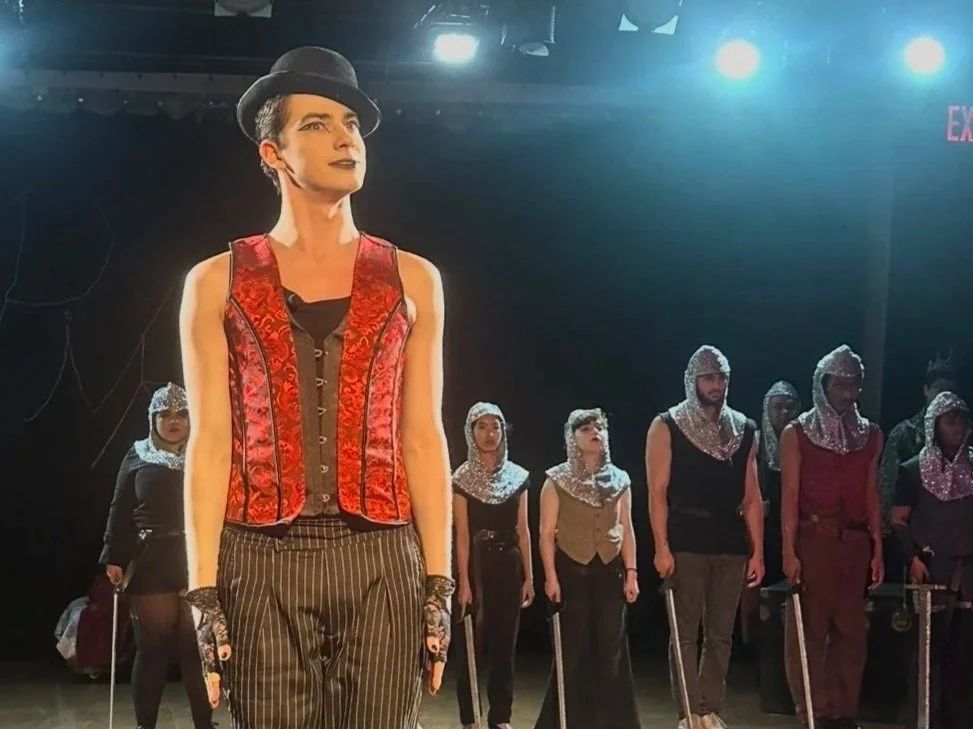 The image shows a stage performance with multiple performers dressed in theatrical costumes, including a woman in the front wearing a red vest, striped pants, a black top, and a gray hat. She has makeup on her face, and other performers behind her we