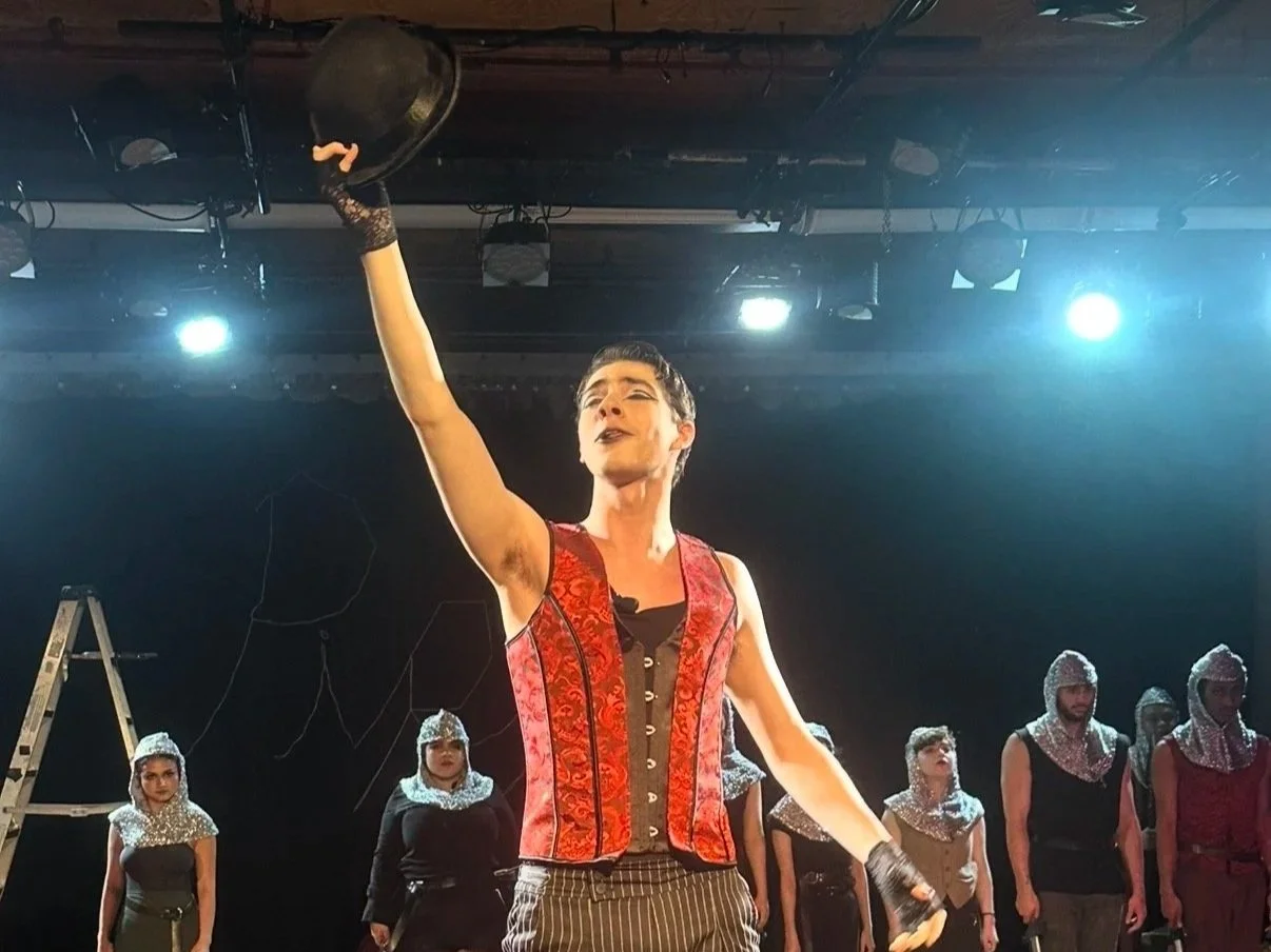 A person dressed in a red and black costume holding a hat high in the air during a stage performance, with several other performers dressed in black with silver head coverings standing behind them on a dark stage.