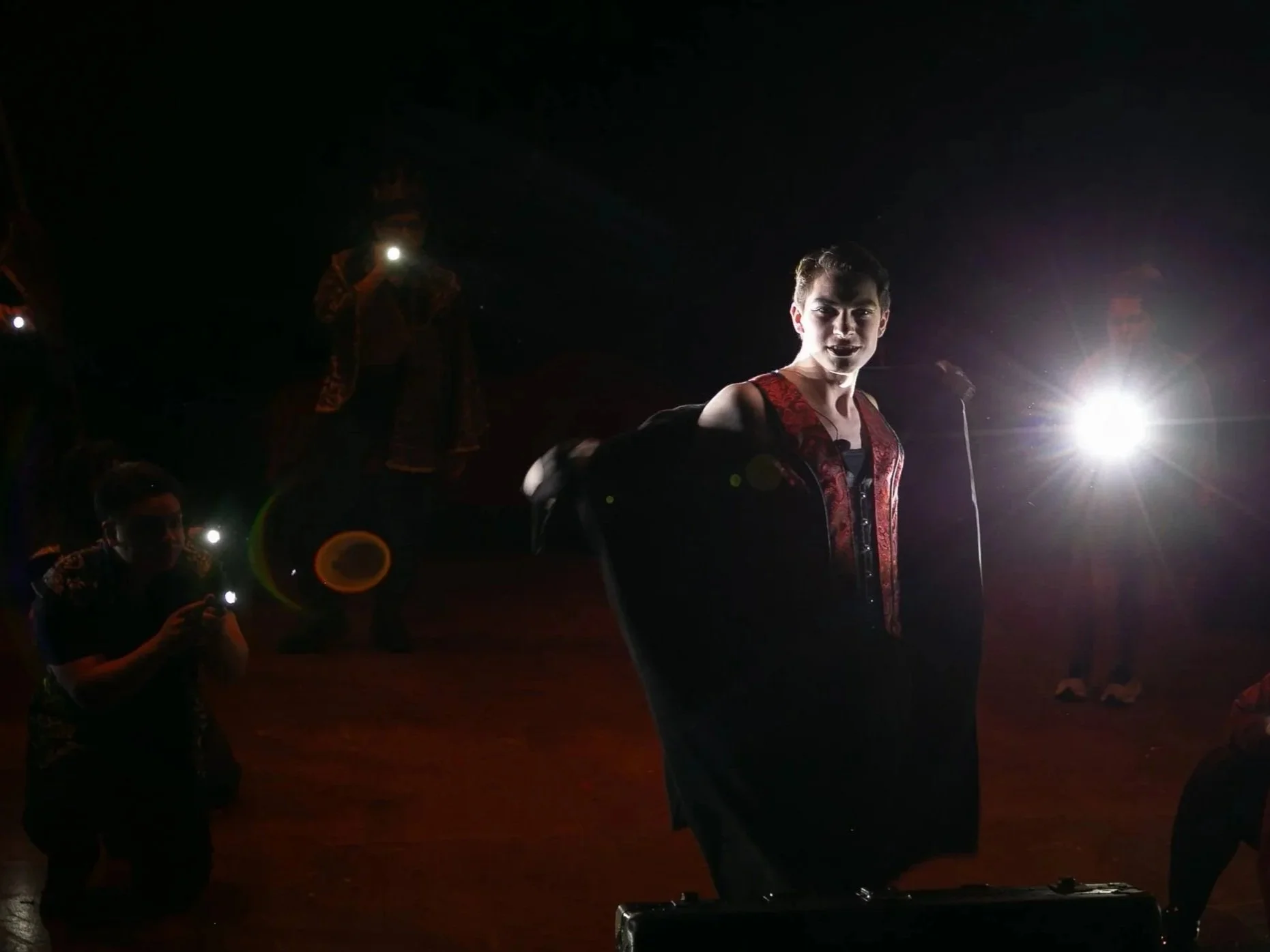 A young man in a dark setting with bright backlighting, wearing a black jacket with red embroidery, looking at the camera. Several people are around, some using their phones or flashlights.