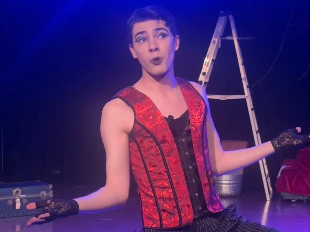 Person wearing a red and black sleeveless vest with lace gloves, performing on stage with a ladder and other equipment in the background.