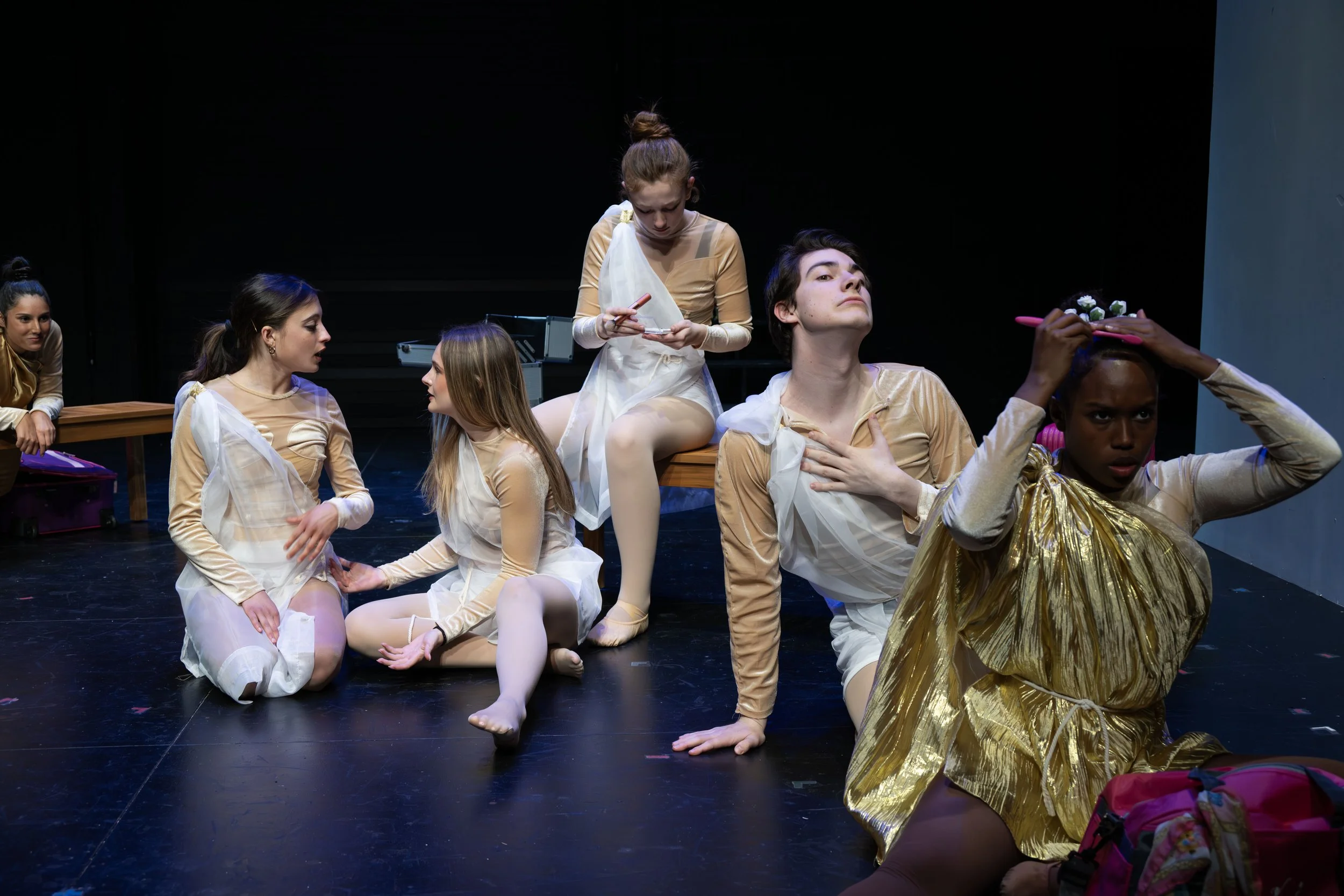 Young performers in gold and white costumes, some sitting and some standing, preparing backstage for a dance recital with makeup, flower accessories, and stage props.
