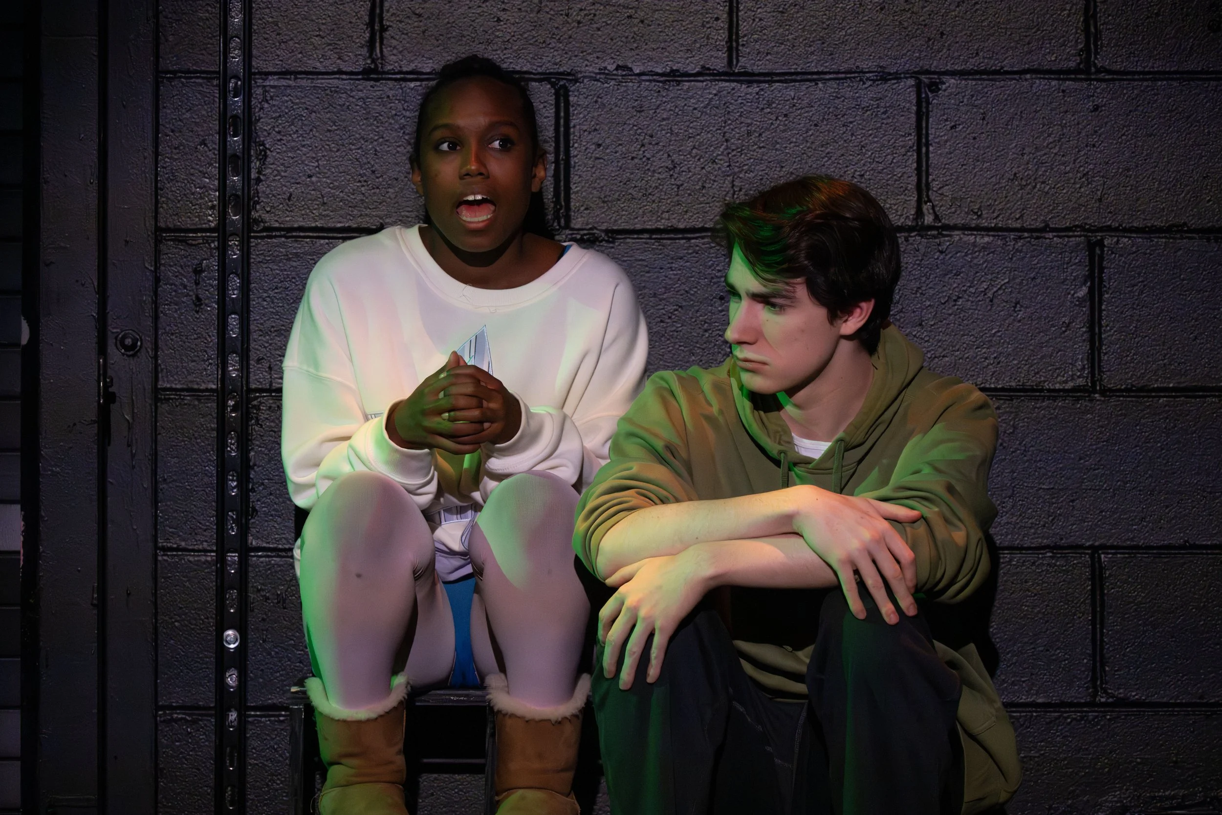Two young people, a woman and a man, sitting against a dark brick wall, with the woman speaking and the man listening. The woman has dark skin, hair tied back, and is wearing a light-colored sweatshirt. The man has light skin, dark hair, and is weari