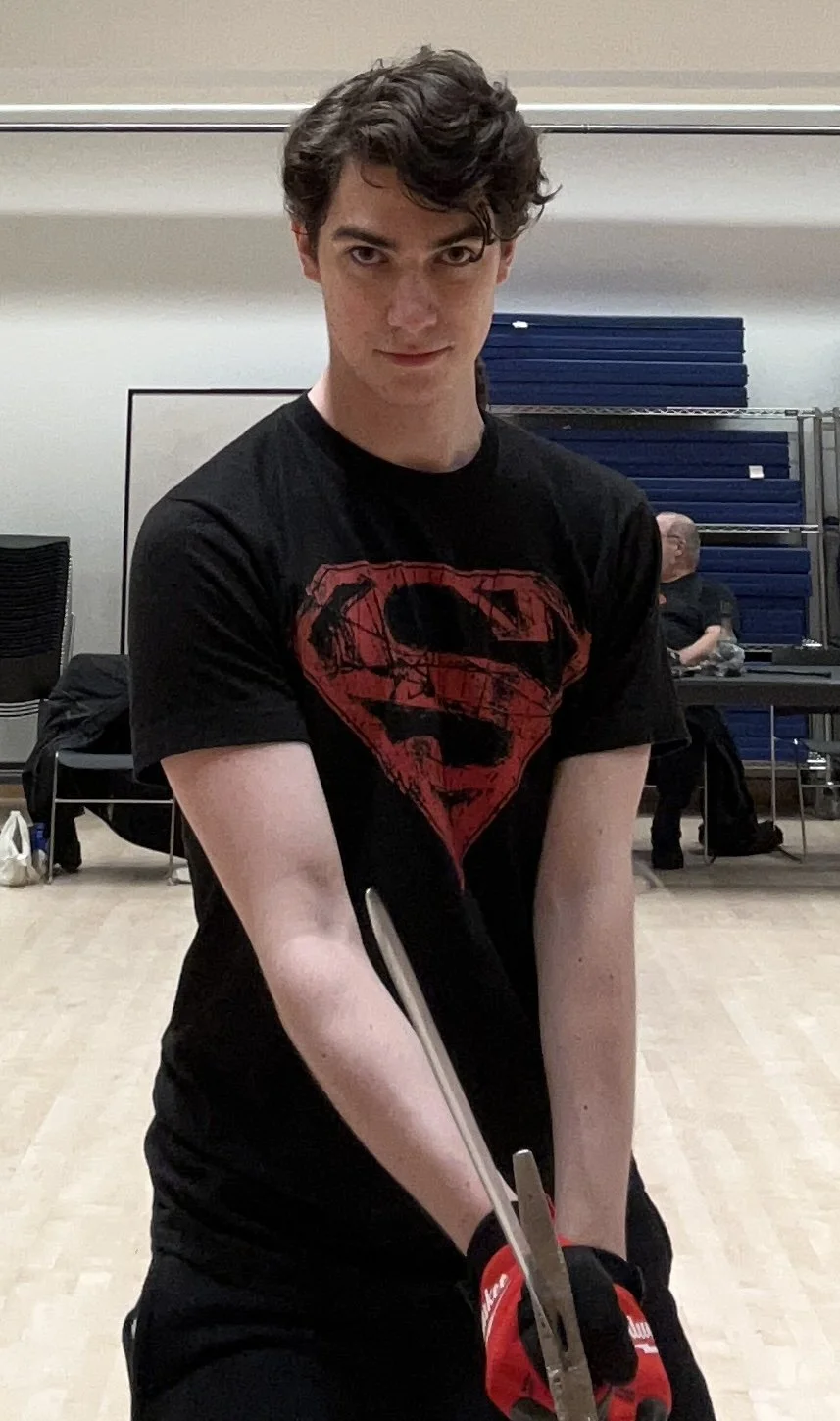 Young man with curly brown hair wearing a black T-shirt with a red Superman logo, holding a sword, in a room with chairs and tables in the background.