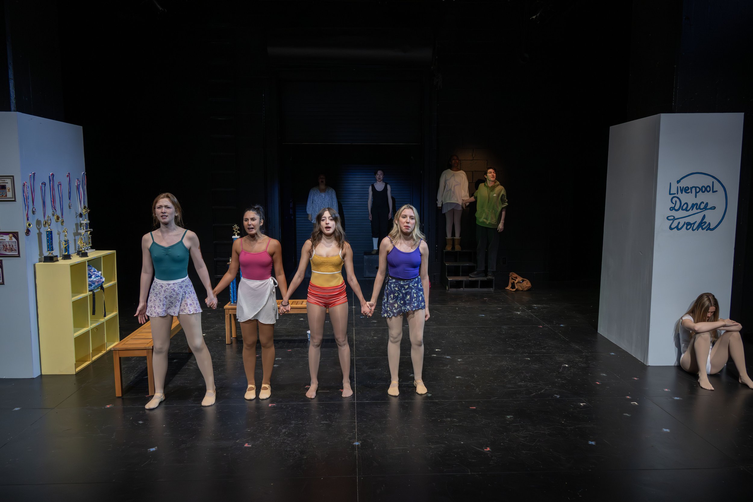 A stage performance with five young women holding hands in colorful shorts and leotards, standing in a line while other performers are visible in the background. There are trophies and medals on yellow shelves to the left and a sign reading 'Liverpoo