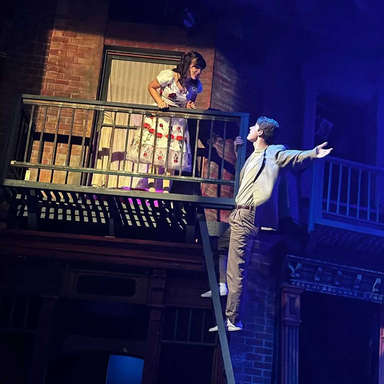 Two actors performing on a theater stage with a brick building facade, one actor standing on a ladder with arms outstretched, and the other actor leaning over a balcony, engaging in a scene.