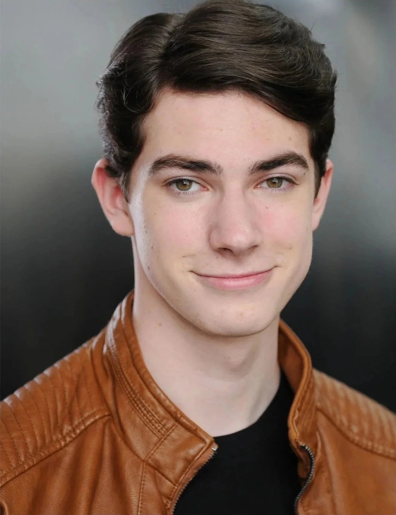 Young man with brown hair and hazel eyes wearing a brown leather jacket and black shirt, smiling slightly, against a plain dark background.