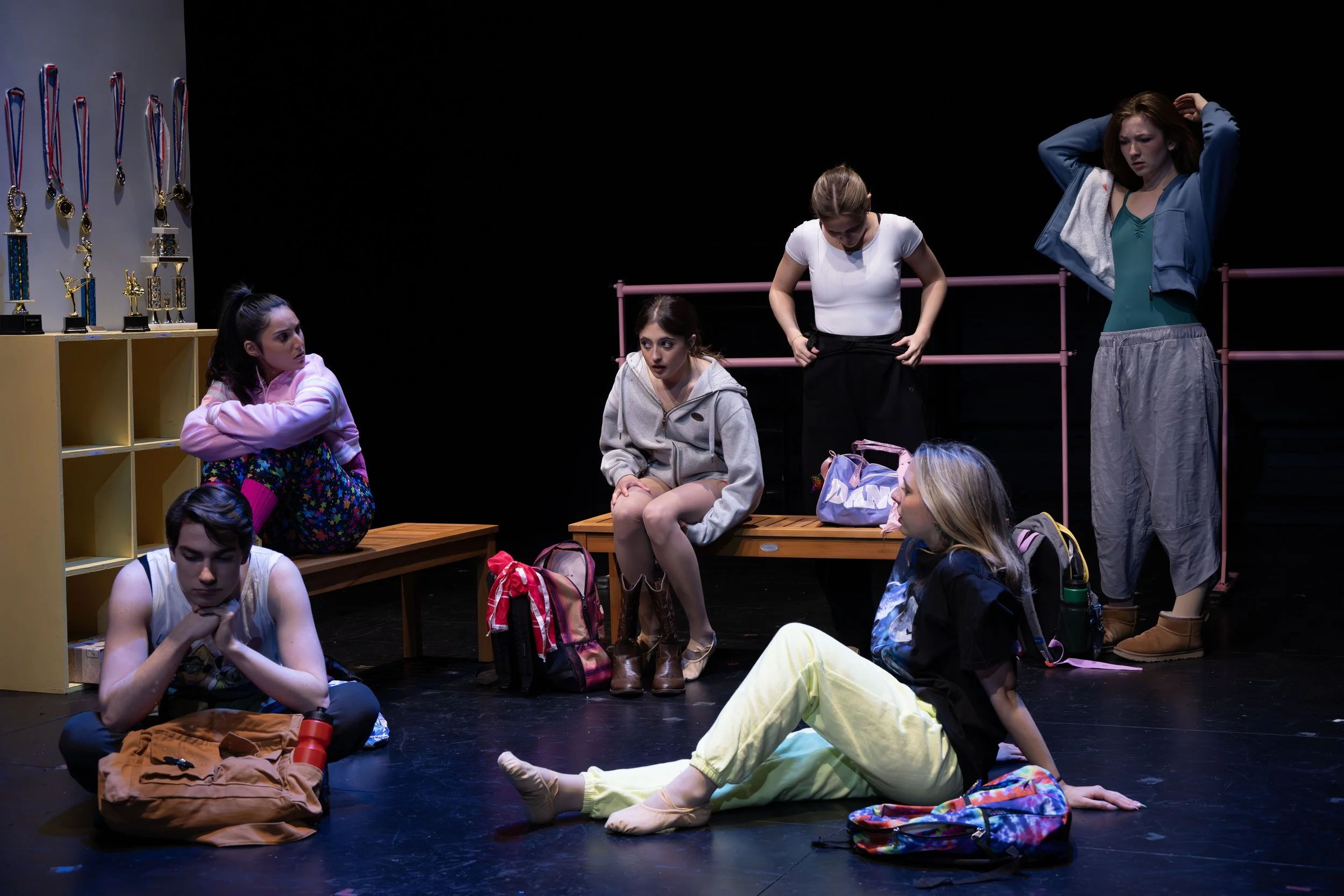 The image shows a group of young women backstage or in a rehearsal space, preparing for a performance. They are sitting and standing amidst personal belongings, with a display of medals on the wall to the left.
