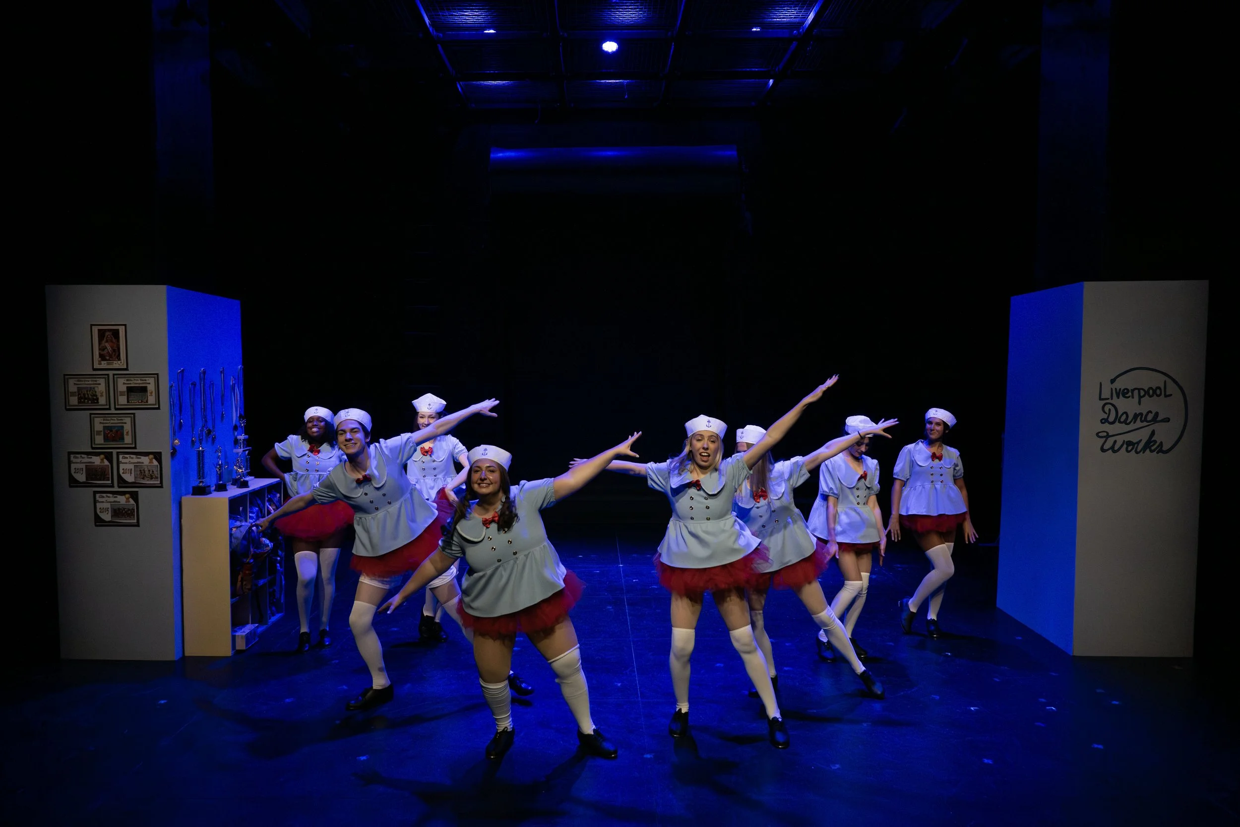 Group of performers in costume dancing on stage as part of a show, with stage decorations including framed photos and signs reading 'Liverpool Dance Works'.