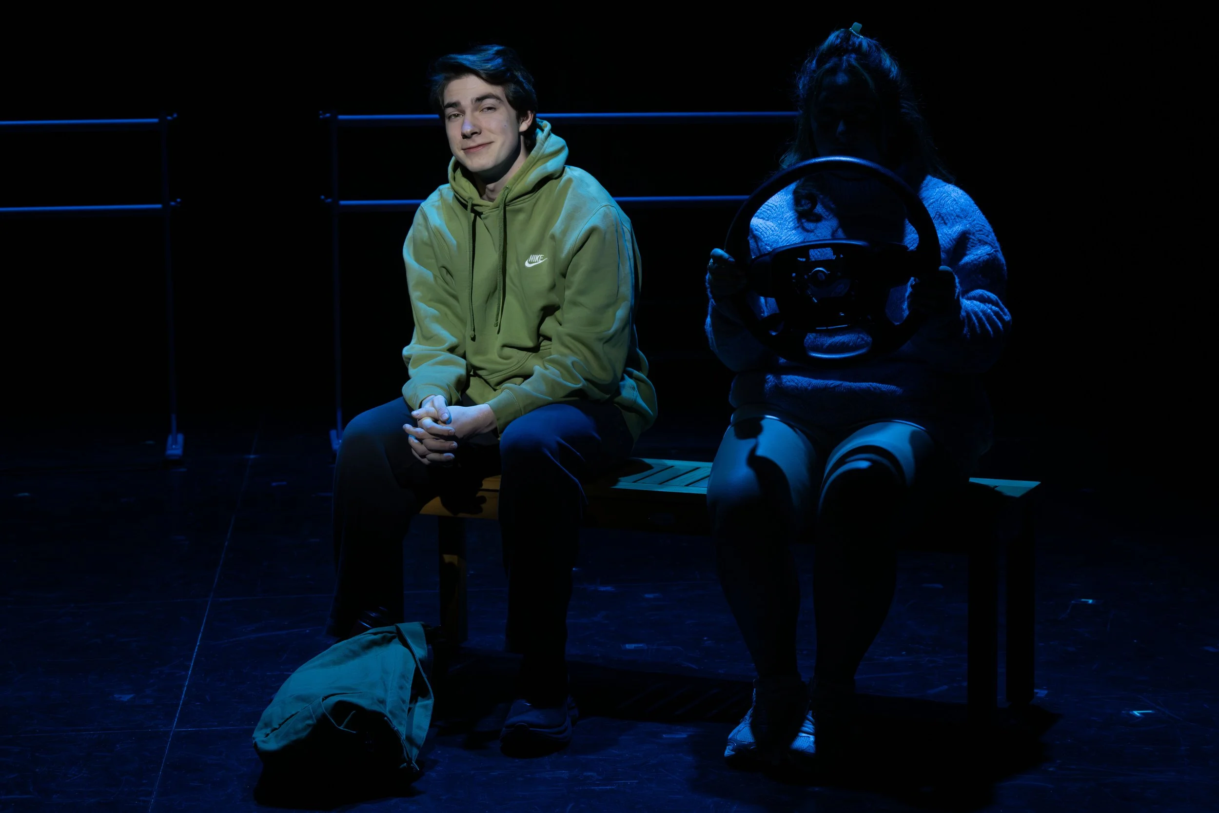 A young man in a green Nike hoodie sitting on a bench, smiling, with a woman to his right holding a steering wheel, both in a dark stage setting with minimal blue lighting.