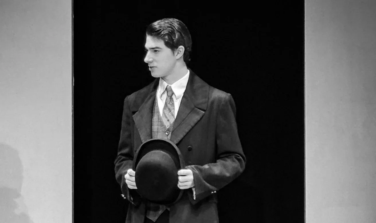 Black and white photo of a young man in vintage clothing holding a bowler hat, standing against a dark background.
