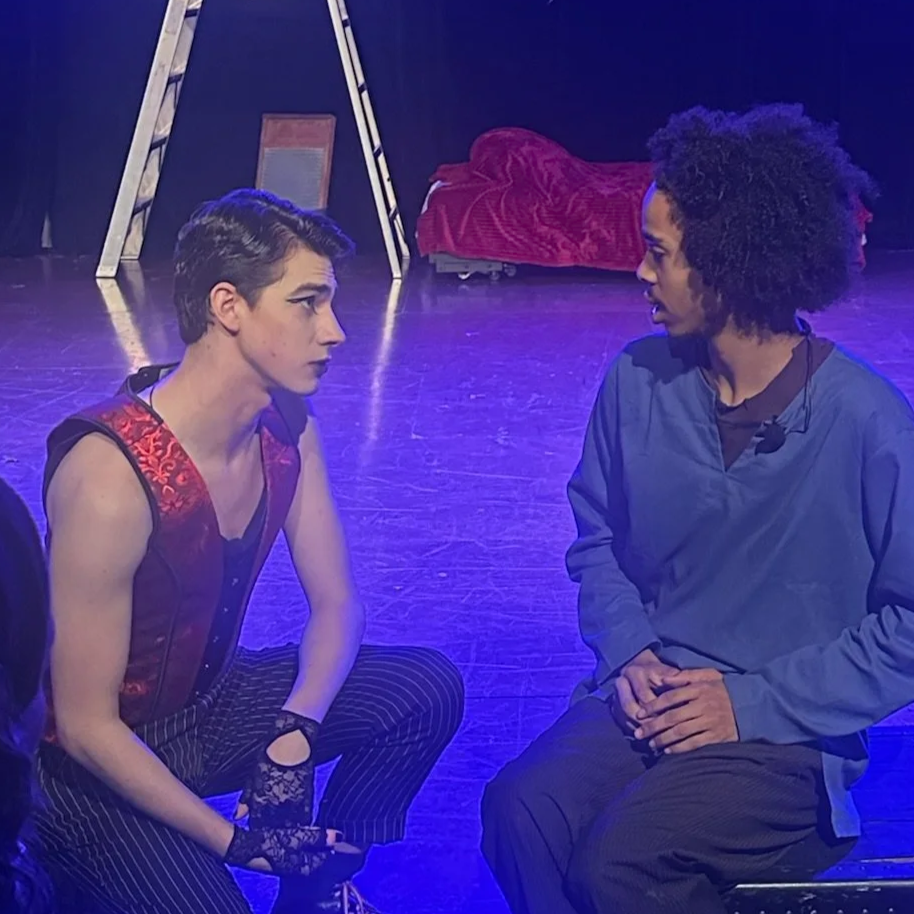 Two performers kneeling and facing each other during a theatrical performance. The person on the left has short dark hair, a sleeveless top with a patterned vest, and striped pants. The person on the right has curly hair, wears a blue shirt and black pants. There is a ladder, a mirror, and a red blanket in the background on a stage with blue lighting.