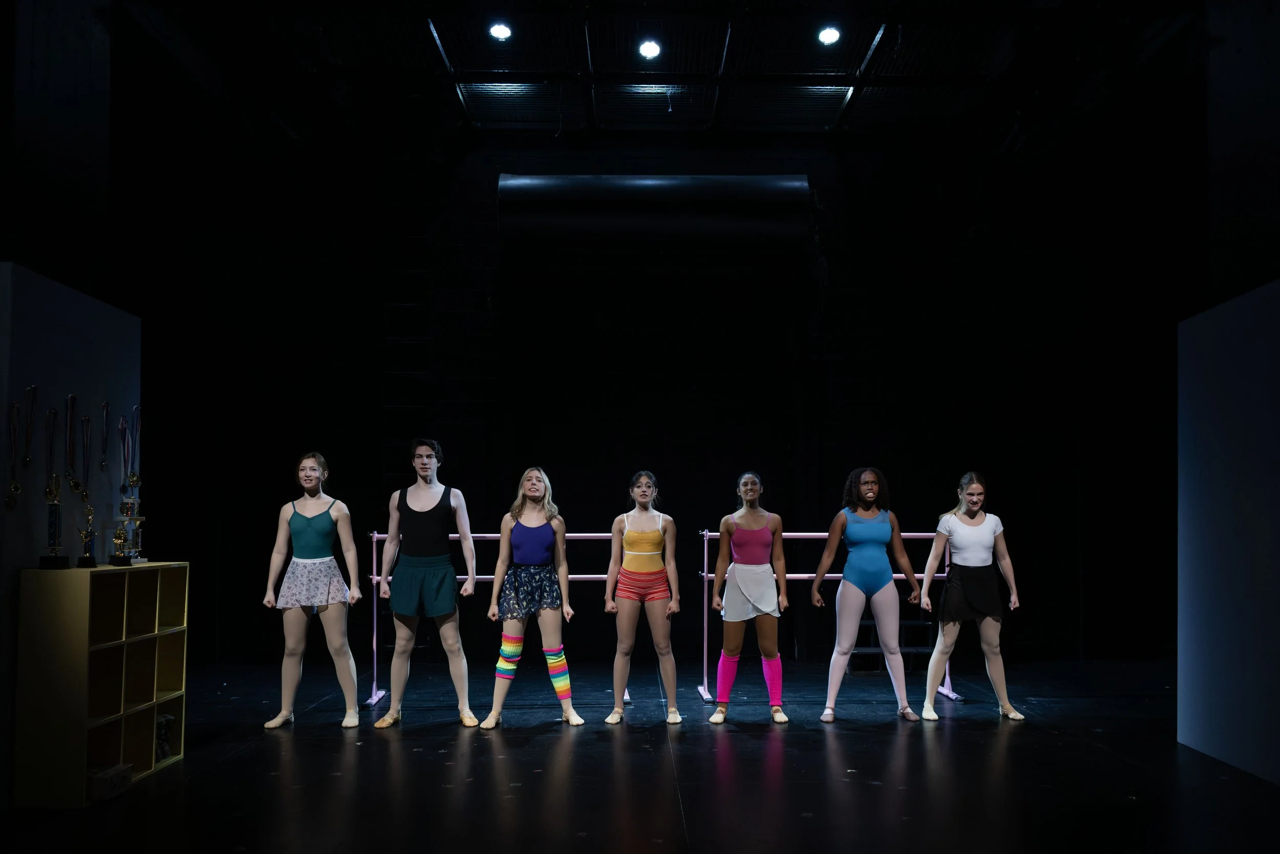 A group of diverse women in colorful dance or ballet attire standing on stage with hands behind their backs, set against a black background with stage lights.