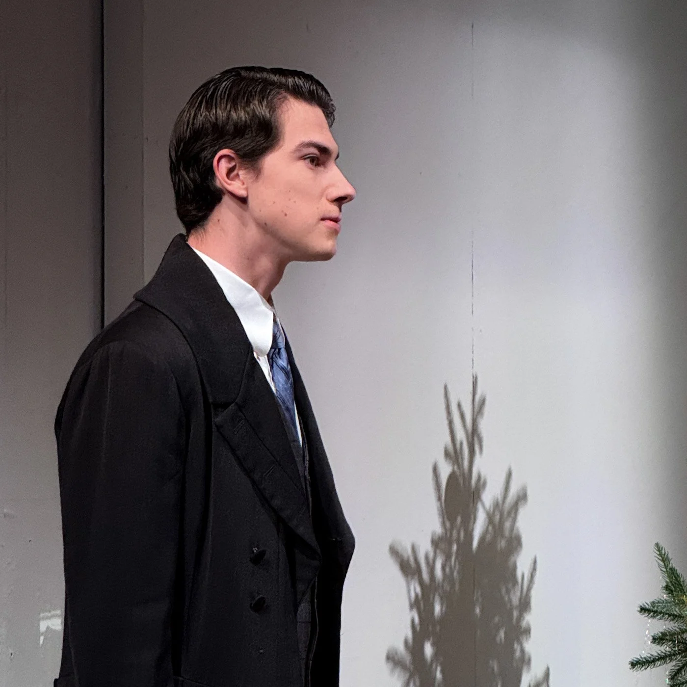 A young man in a black coat and tie stands against a plain wall, with a shadow of a Christmas tree cast on the wall beside him.