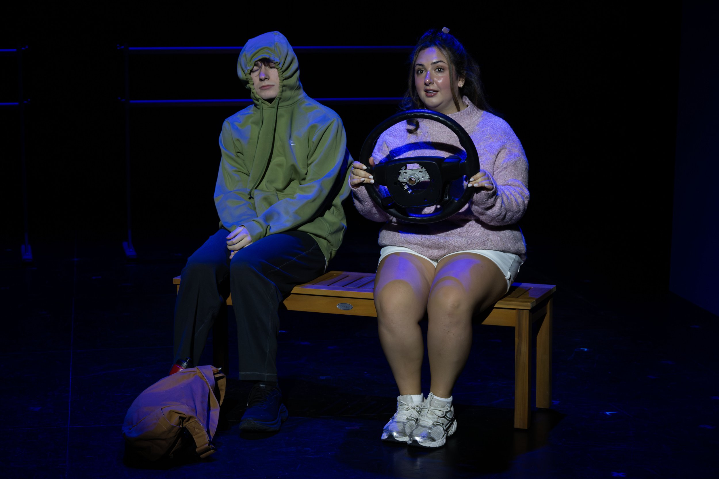 Two young women sitting on a wooden bench on a dark stage. The woman on the left is wearing a green hoodie with the hood up and appears to be sleeping or resting her eyes. The woman on the right is holding a steering wheel prop, wearing a pink sweate