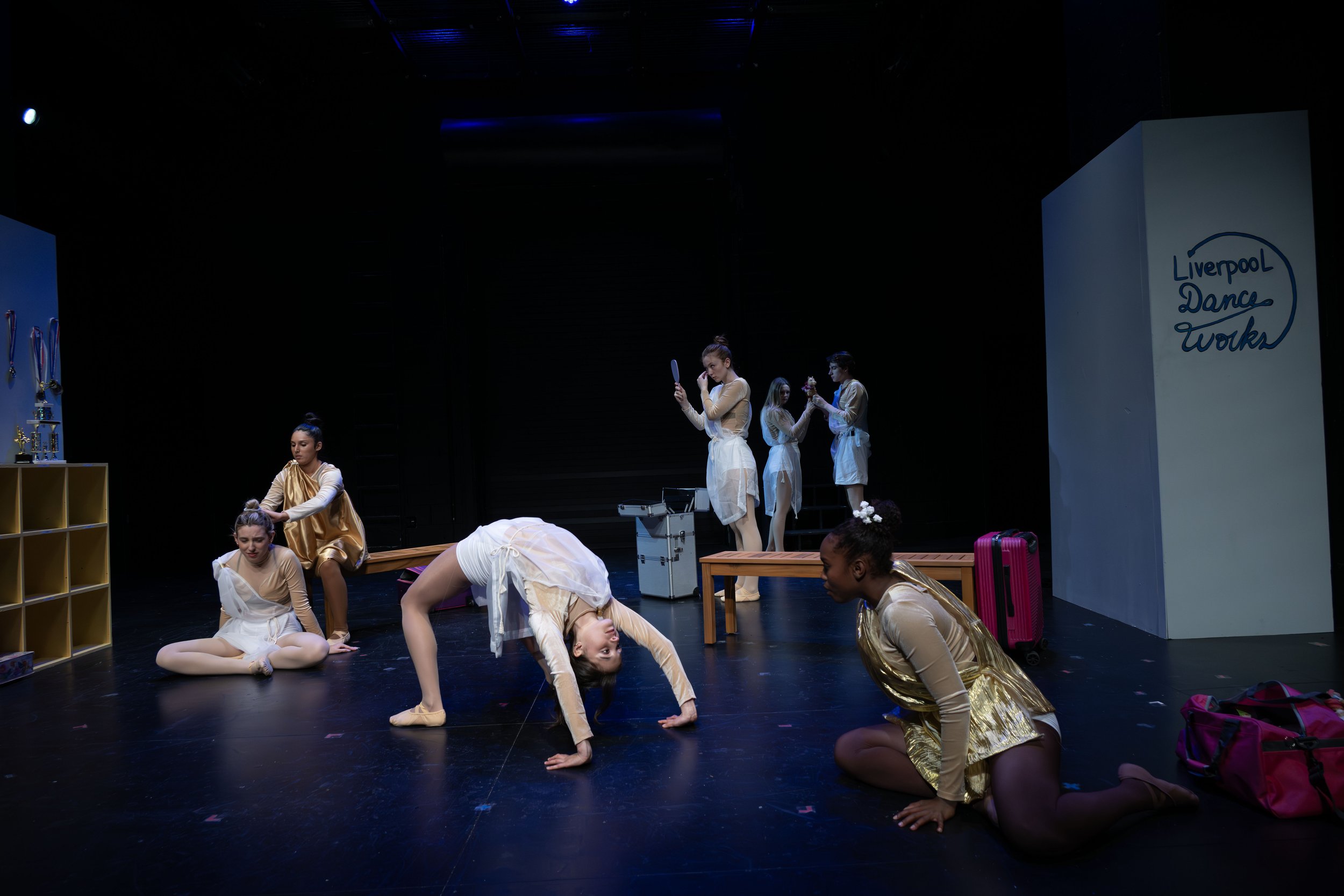 A group of young dancers in beige and gold costumes practicing on stage. Some are stretching or doing yoga poses, while others are standing, sitting, or lying on the floor. Stage props include shelves, a pink suitcase, as well as a large white pillar