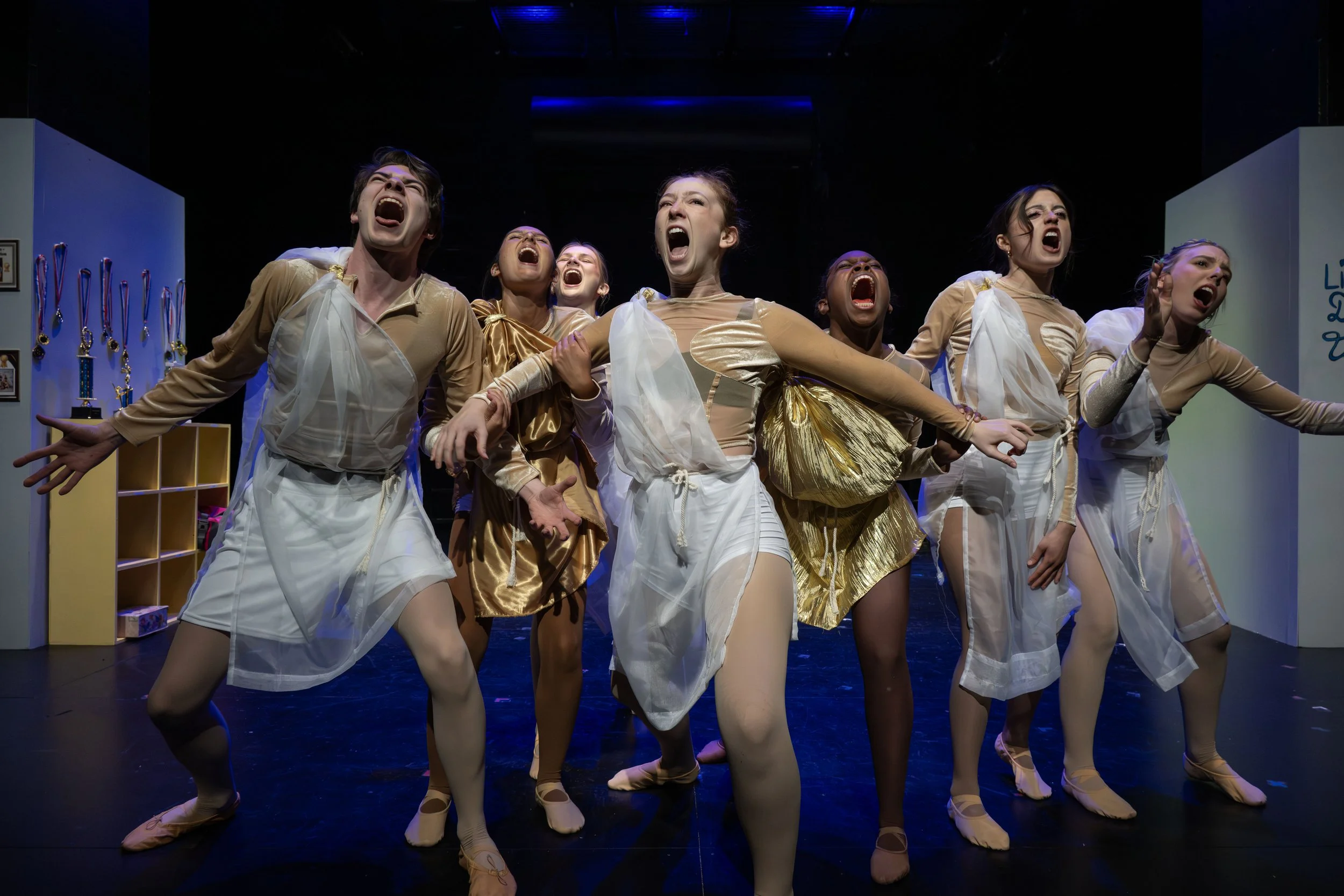 Group of dancers performing on stage with animated expressions, wearing coordinated costume outfits in beige, white, and gold, with some holding hands and displaying energetic movements.