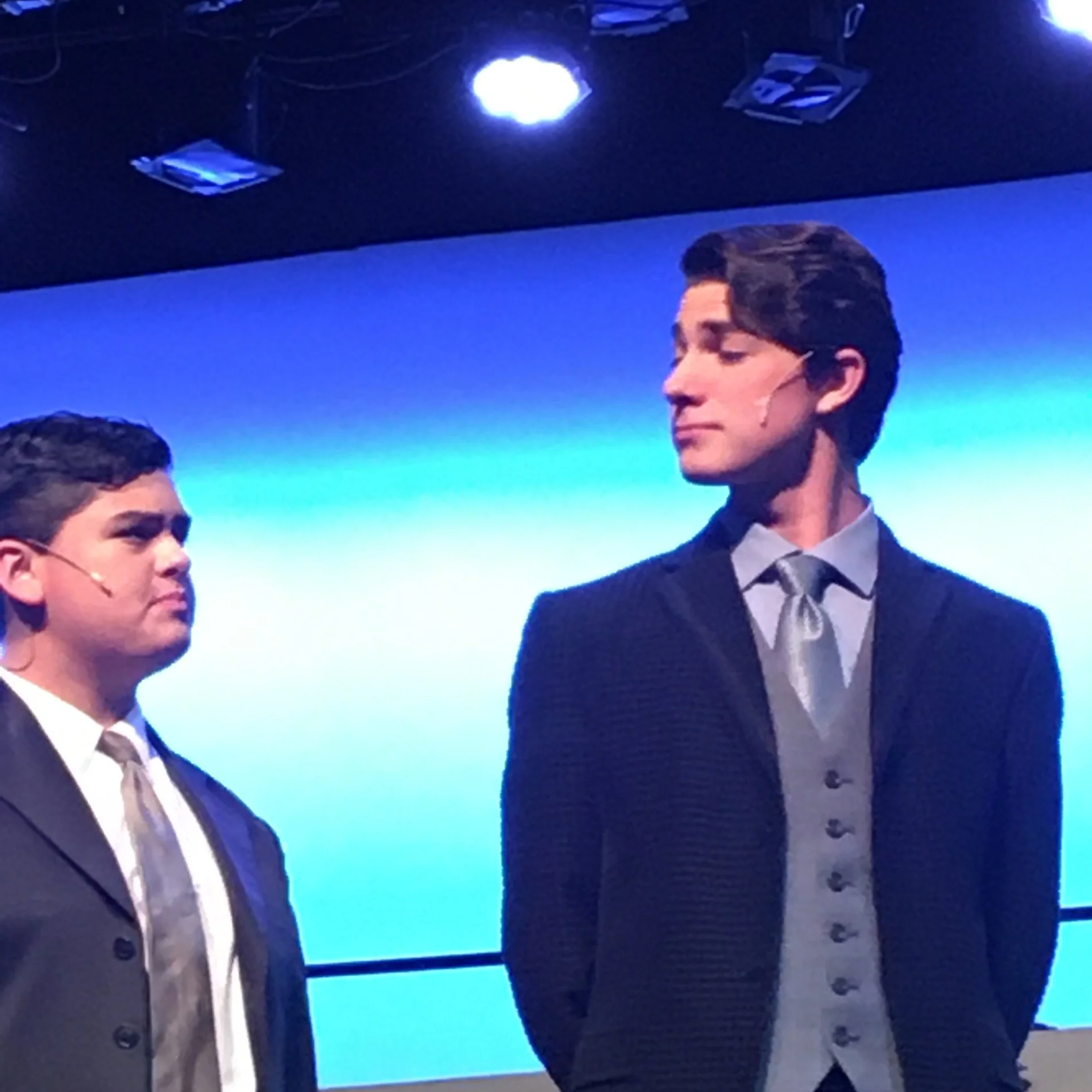 Two men in suits and glasses standing on a stage with a blue background, with ceiling lights and equipment visible.