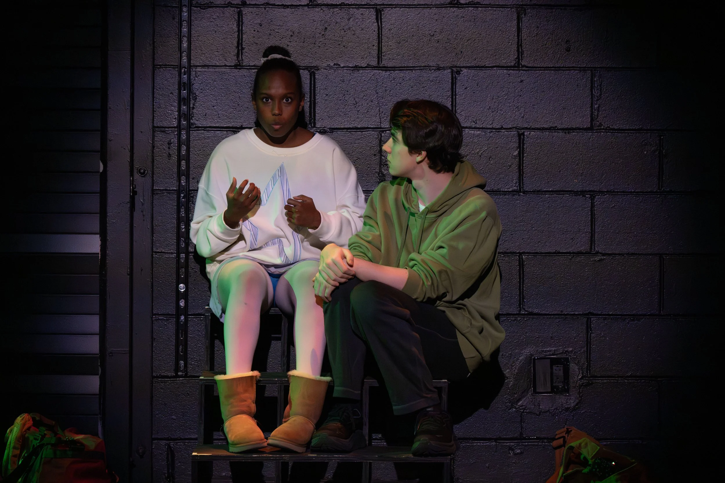 A young woman and man sitting against a black brick wall, engaged in conversation. The woman is wearing an oversized white sweatshirt and tan boots, while the man is dressed in a brown hoodie and dark pants. There are some bags and shoes on the floor