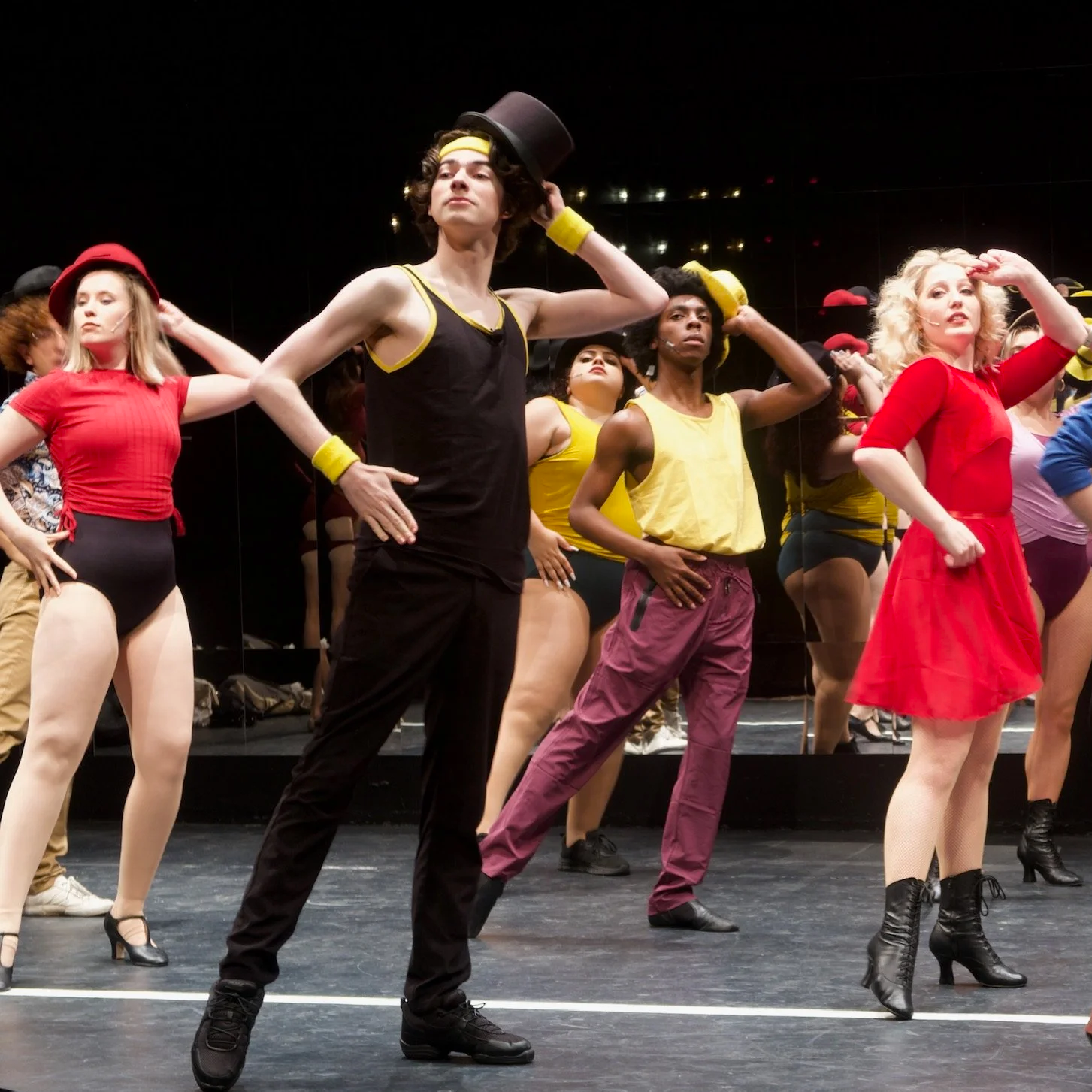 Group of performers on stage in colorful costumes, posing with confident expressions, in a theatrical setting.