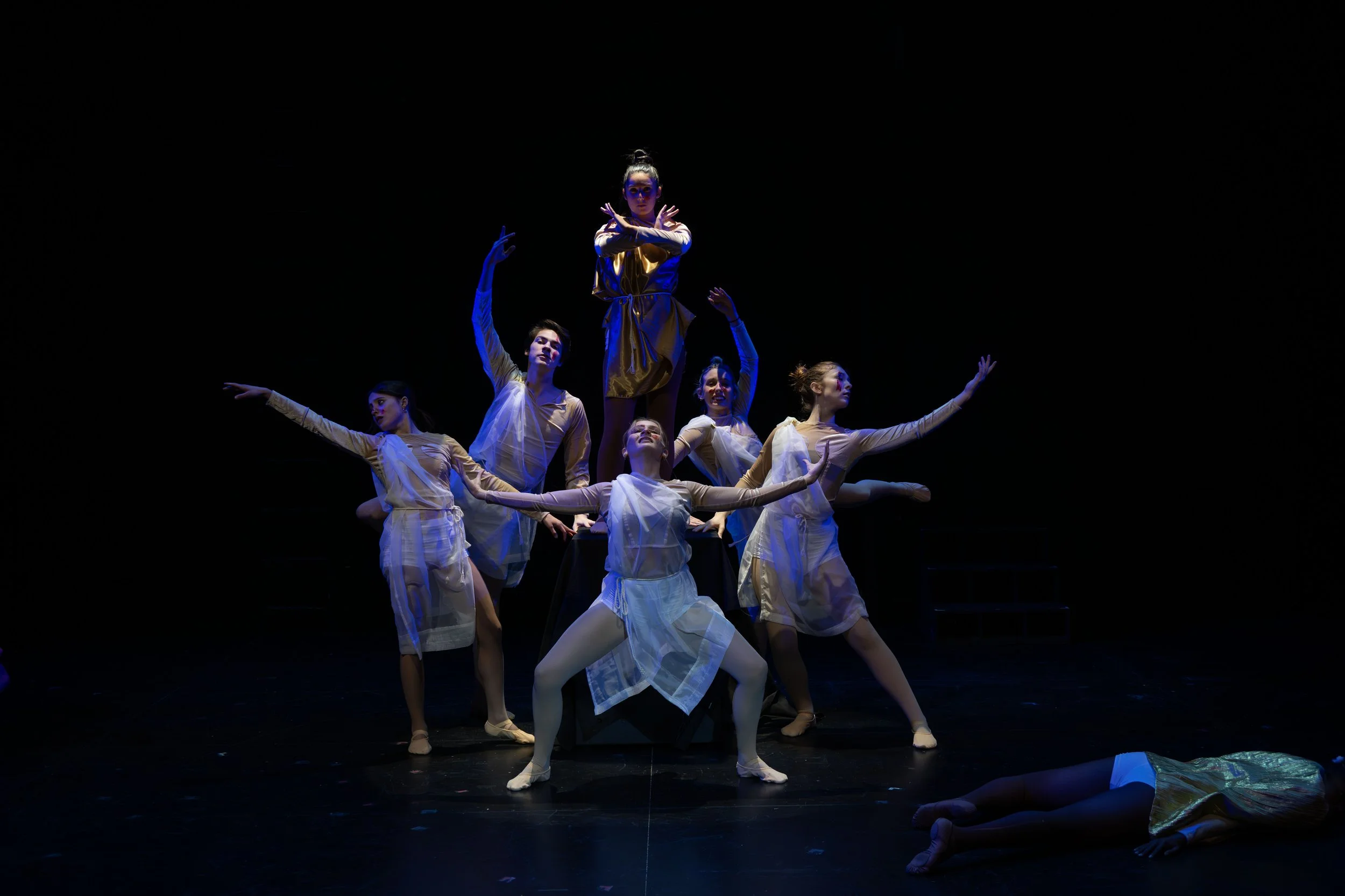 A group of six female ballet dancers performing on a dark stage, with the central dancer elevated and crossing her arms, surrounded by the others in various poses, all wearing matching light-colored costumes.