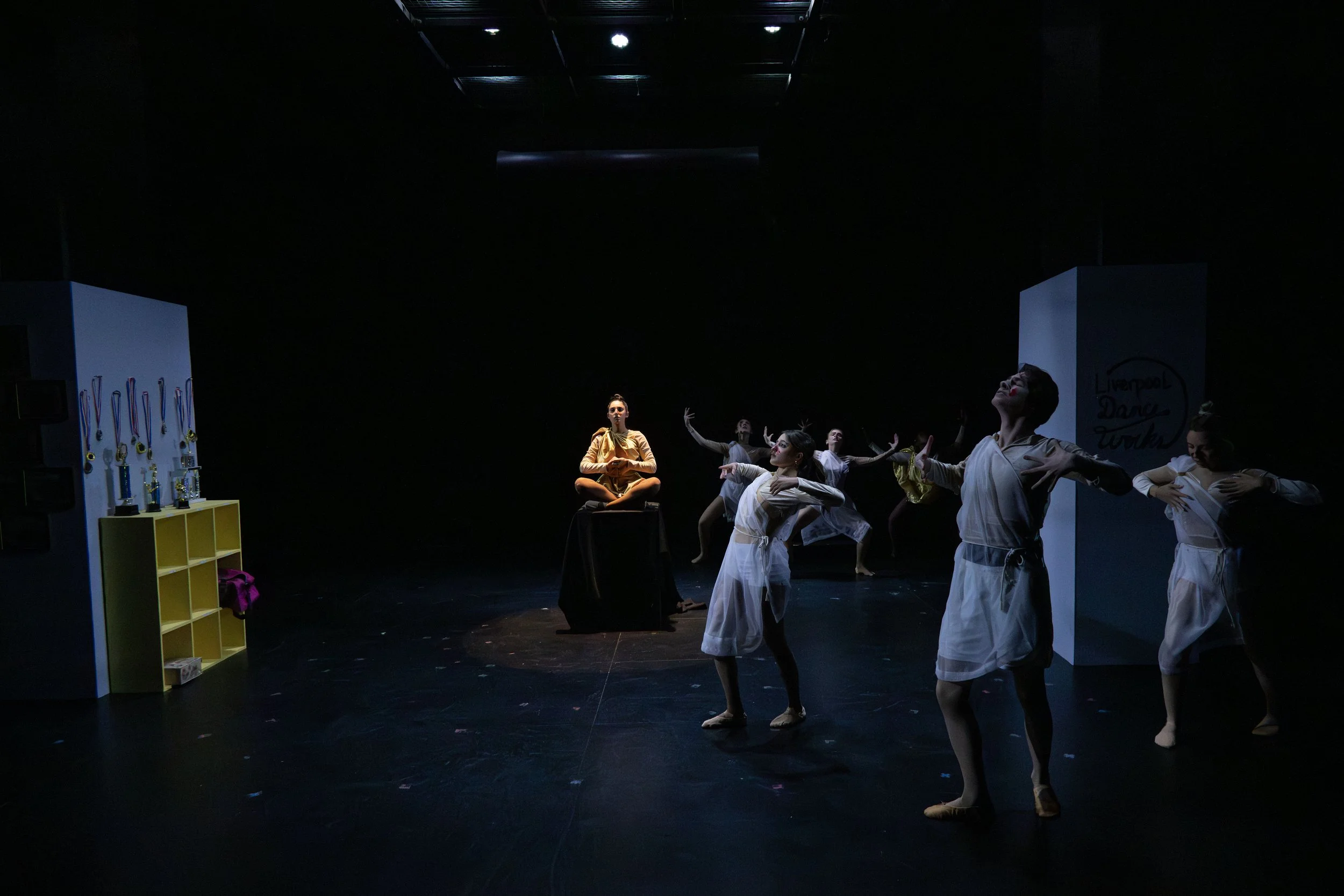 The image shows a stage performance with multiple dancers in white costumes, some with red face paint, in various dance positions. There is a woman sitting cross-legged in the center, illuminated by a spotlight. The background is dark, with minimal l