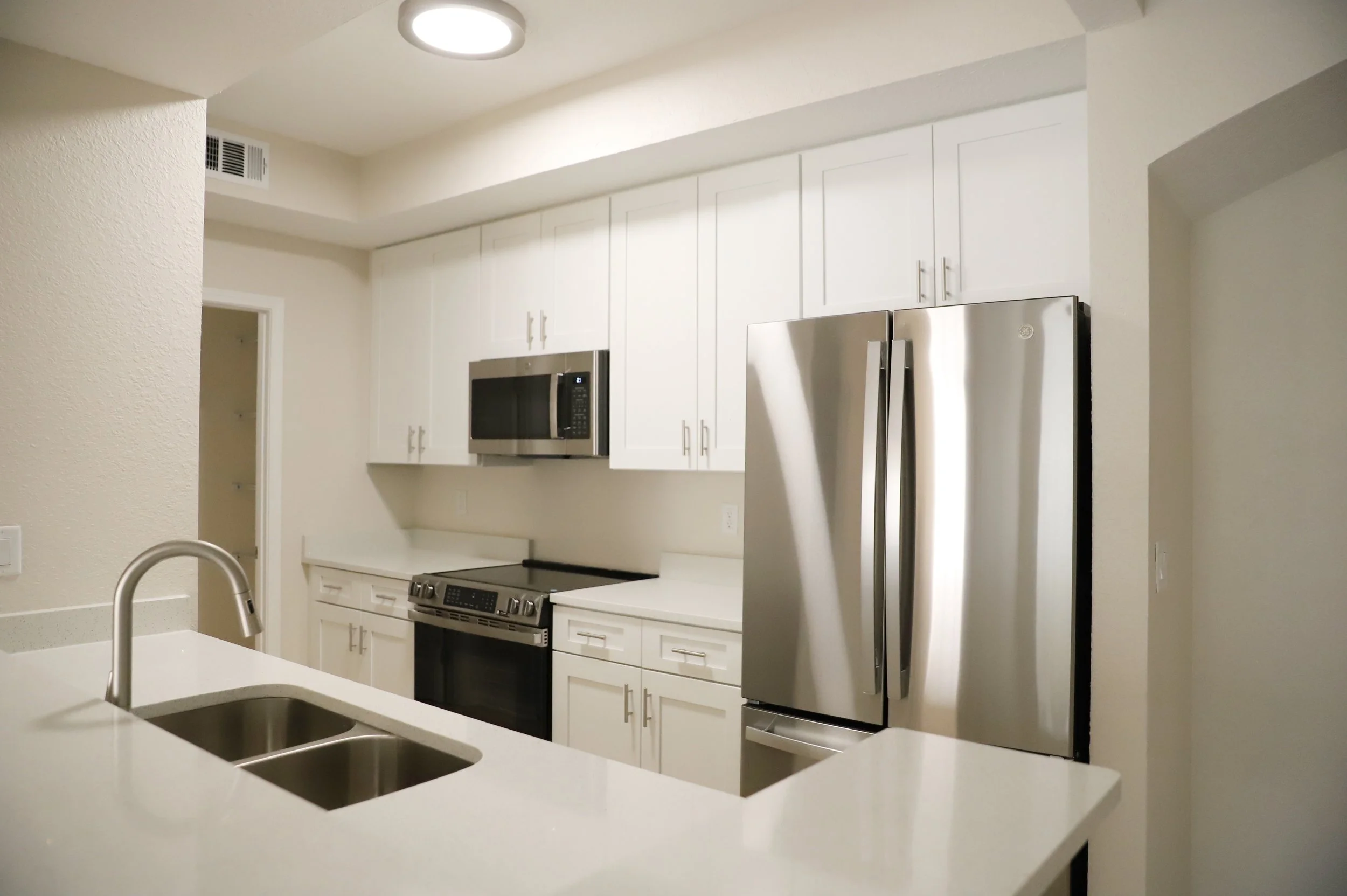Modern kitchen with white cabinets, stainless steel appliances including a refrigerator, microwave, and oven, white countertops, and a double sink with a faucet.