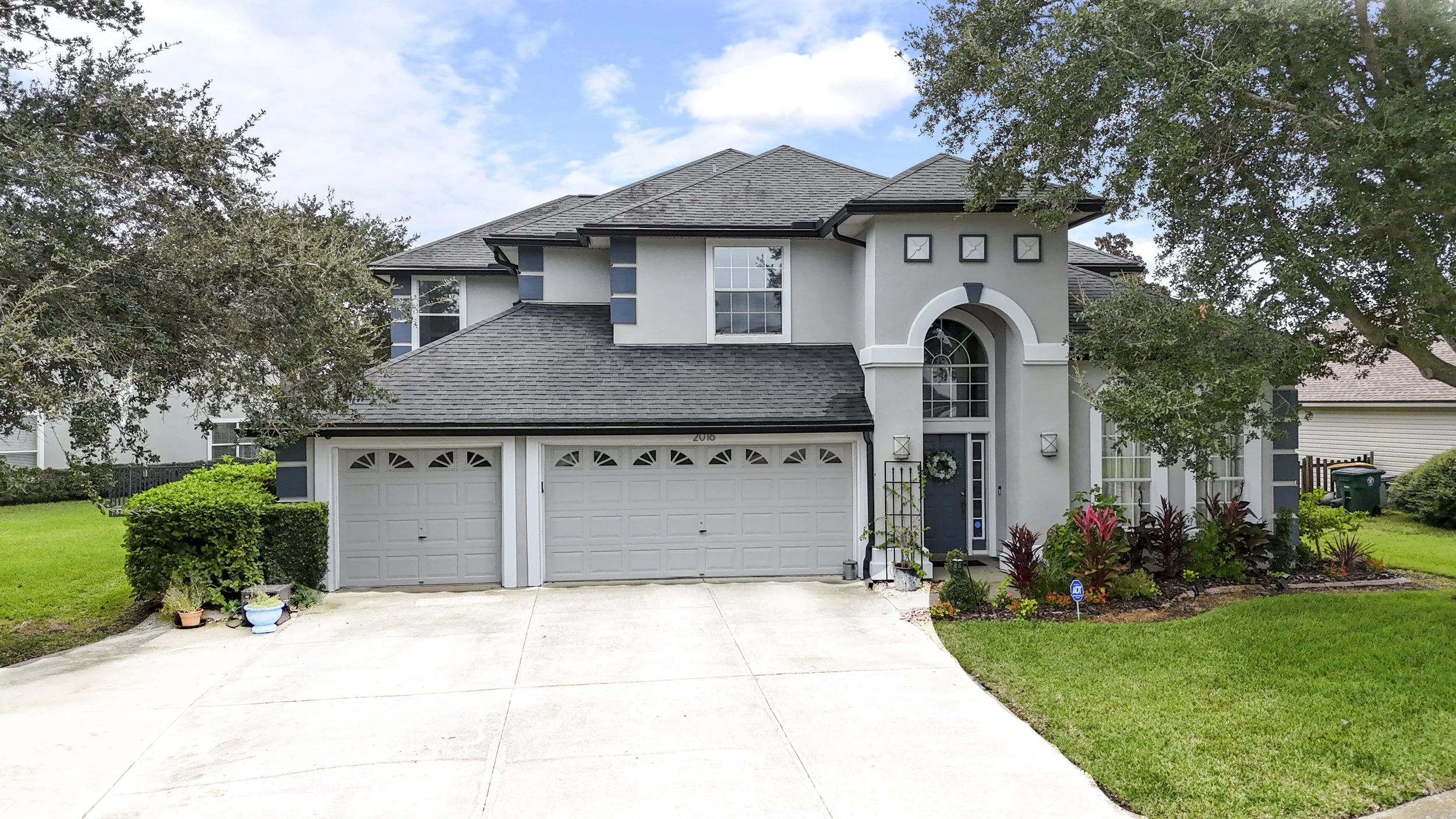 A two-story suburban house with a gray roof, white walls, and a double garage. The front yard has green grass, shrubs, and potted plants, with a driveway leading to the garage. Trees surround the house, and the sky is partly cloudy.