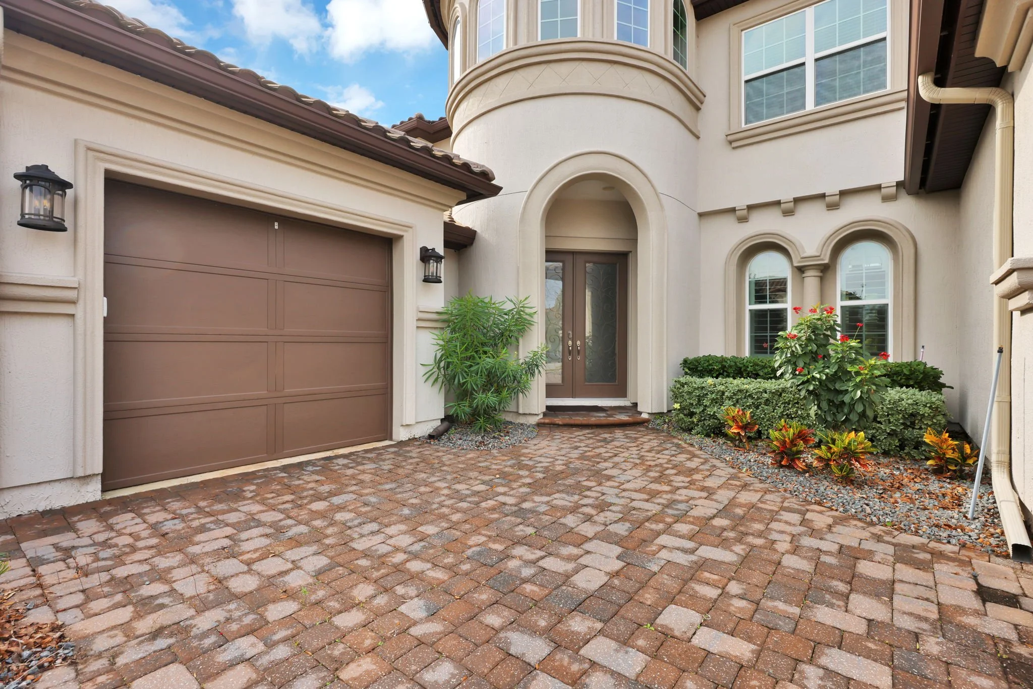 Front entrance of a house with a brown garage door, decorative shrubs, flowers, and a brick driveway.