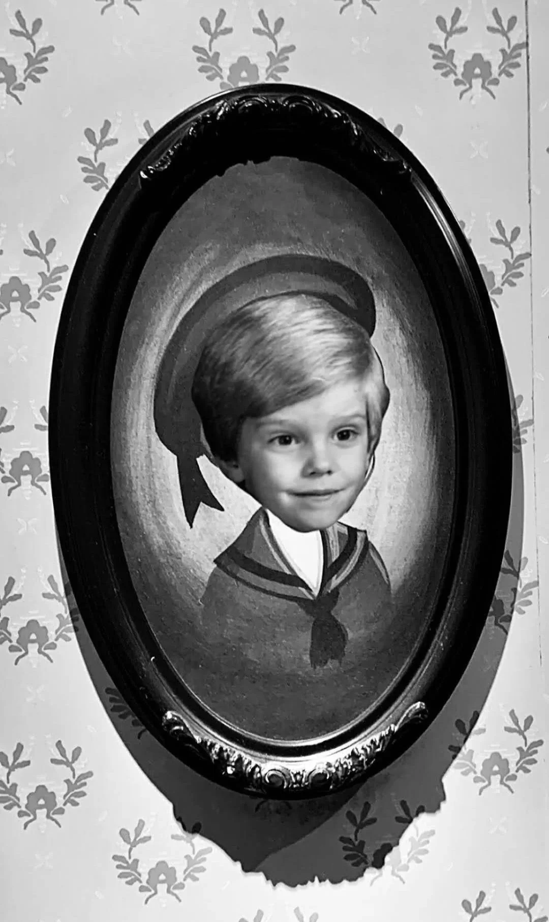 Black and white photo of a vintage oval mirror reflecting a child's face with a superimposed young child's face in the center, resembling a portrait of a boy in a sailor suit.
