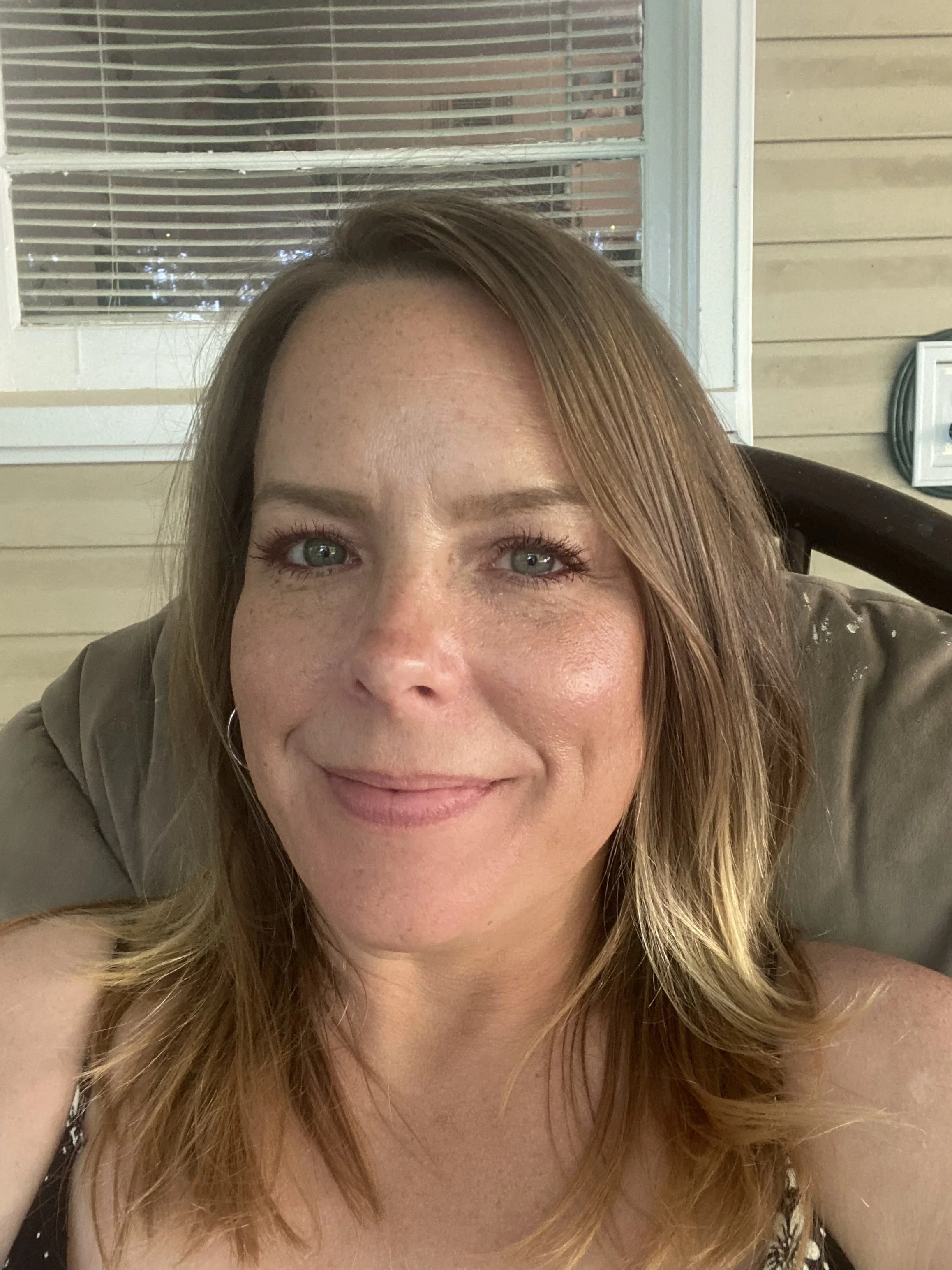 A woman with light skin, freckles, and long, wavy, light brown hair smiling and lying on a surface near a window with blinds and a beige wall behind her.