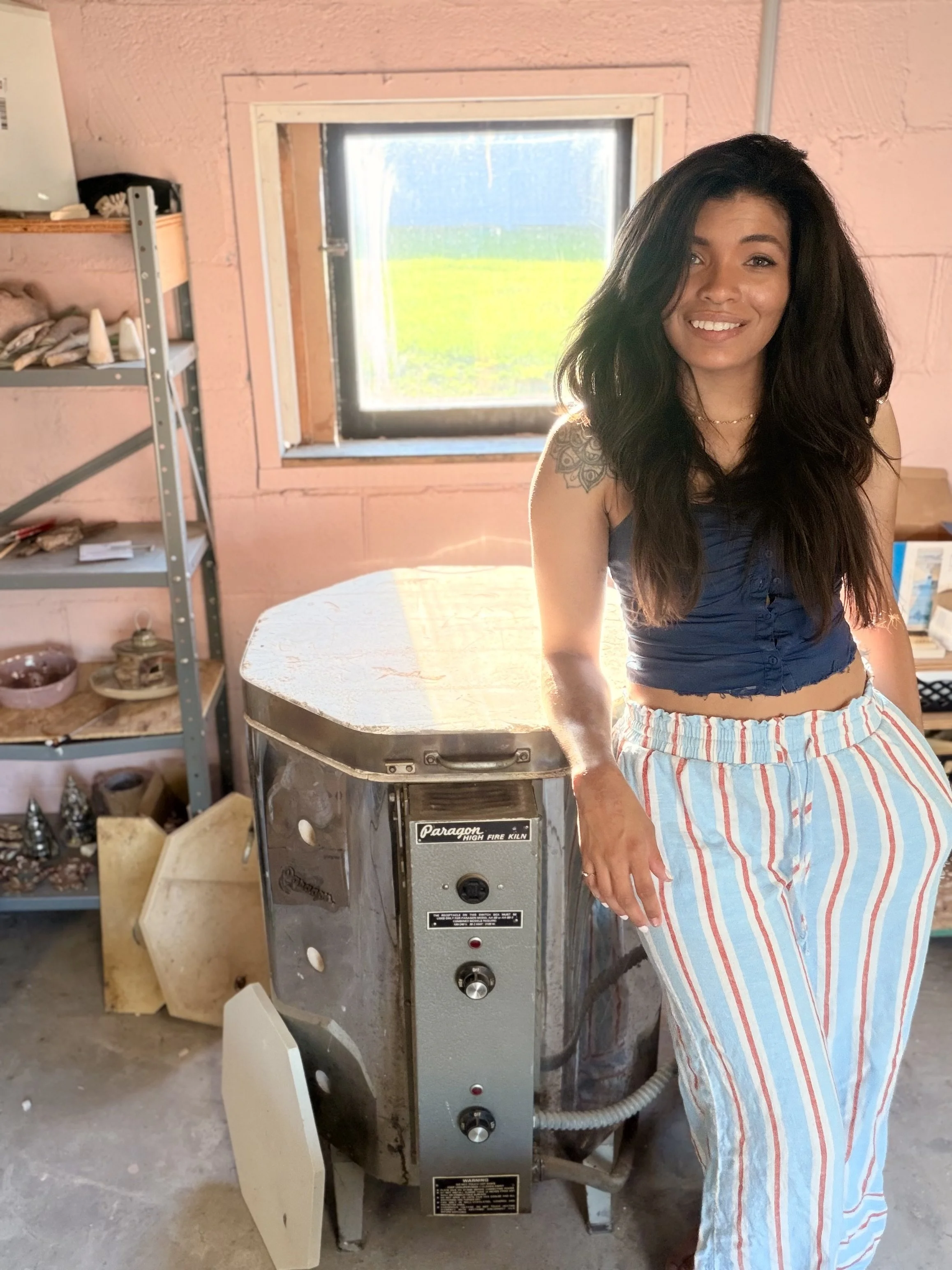 A woman with long dark hair, wearing a blue top and striped pants, standing next to a vintage high fire kiln in a workshop with shelves and various items in the background.