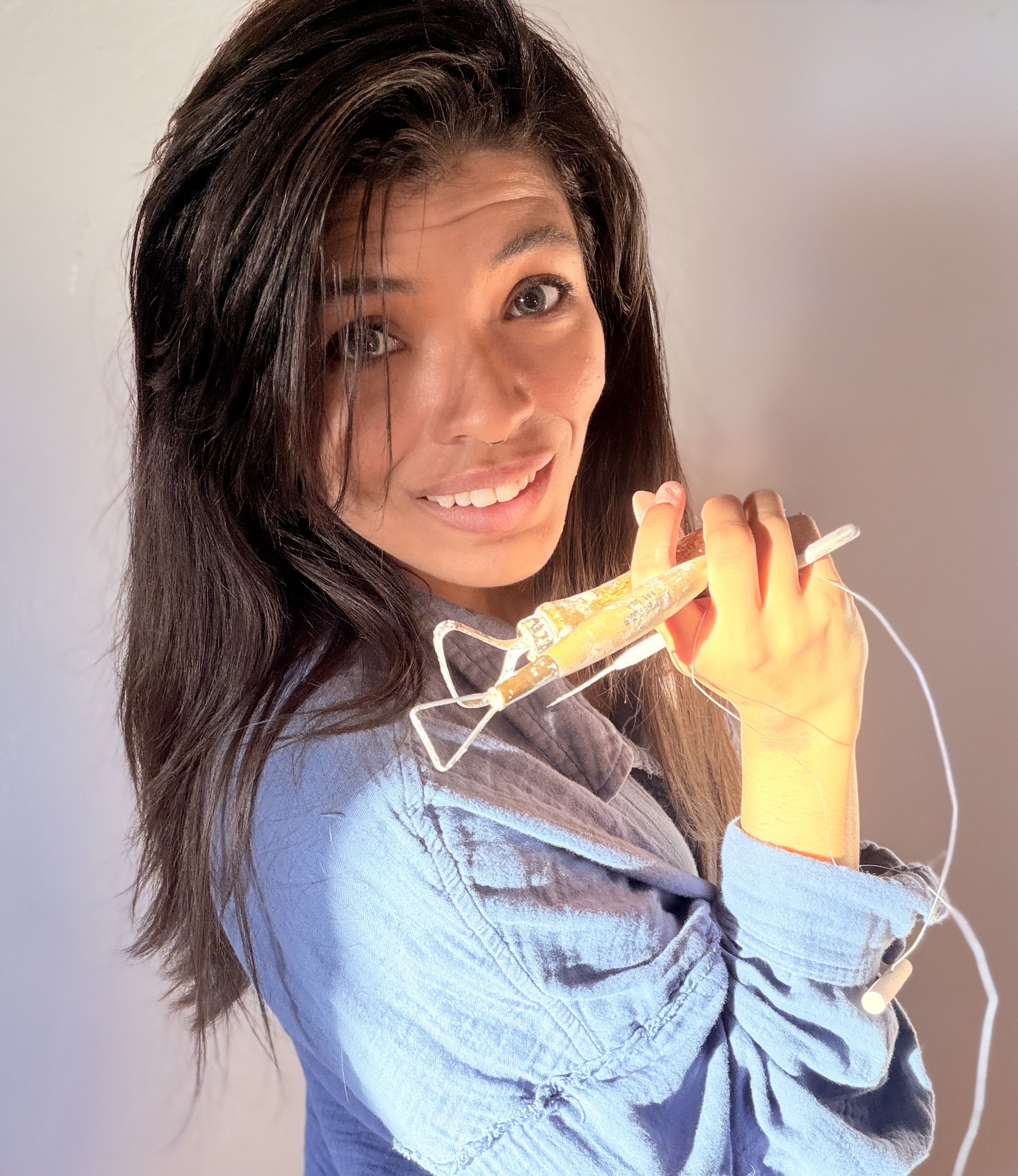 A woman with long dark hair smiling and holding a syringe with a yellow liquid, wearing a denim shirt.