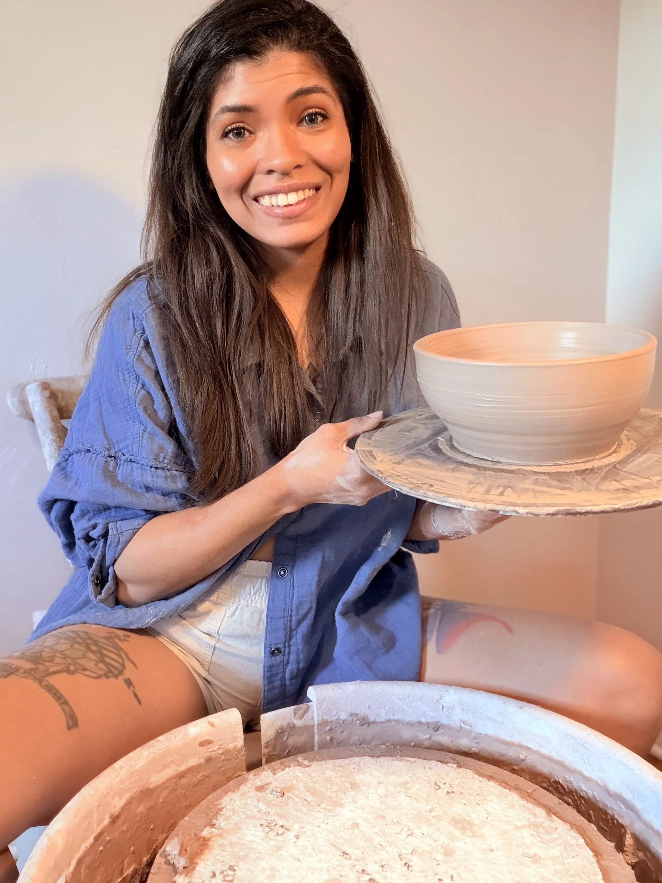 A woman with long brown hair, smiling, holding a pottery wheel with a partially shaped clay bowl, in a pottery studio.