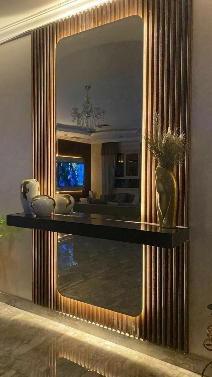 Decorative wall mirror with wooden slats and backlighting, displaying ceramic vases and a tall decorative plant on a black floating shelf in a modern living room.