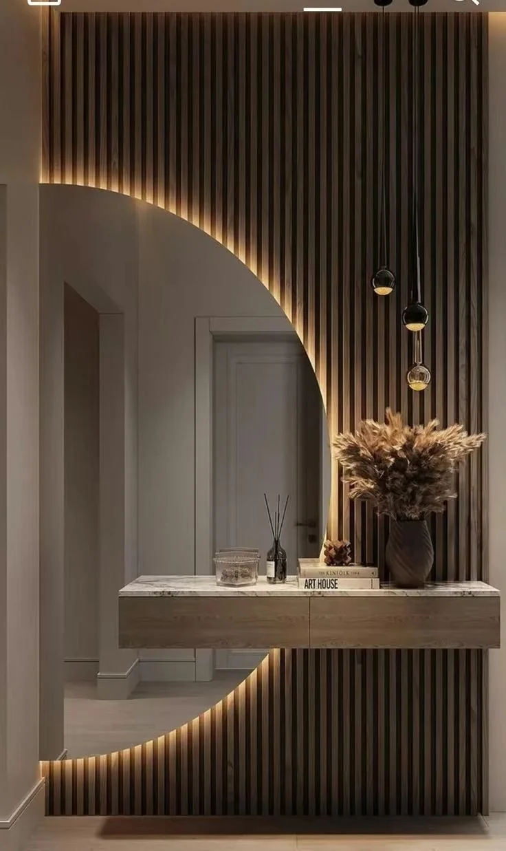 Modern interior wall with vertical wooden slats, a large round mirror with backlit LED lighting, a marble-topped console table with decorative items, including a vase with pampas grass, books, and a diffuser.