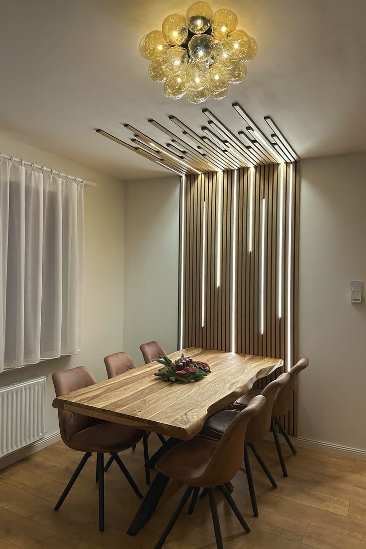 Modern dining room with a wooden table, six upholstered chairs, a decorative centerpiece, wooden panel accents with lighting on the wall, white curtains, and a ceiling light fixture with glass globes.