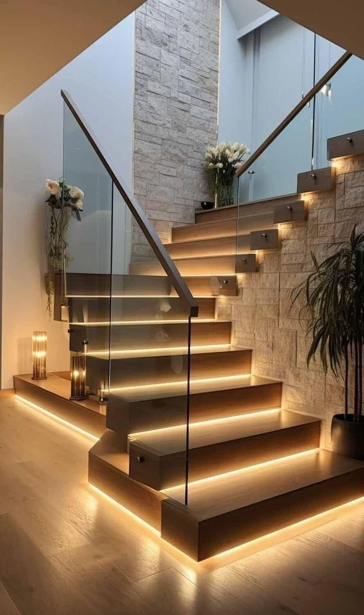 Modern indoor staircase with wooden steps, glass railing, and LED lighting underneath each step, decorated with flower arrangements and a potted plant.