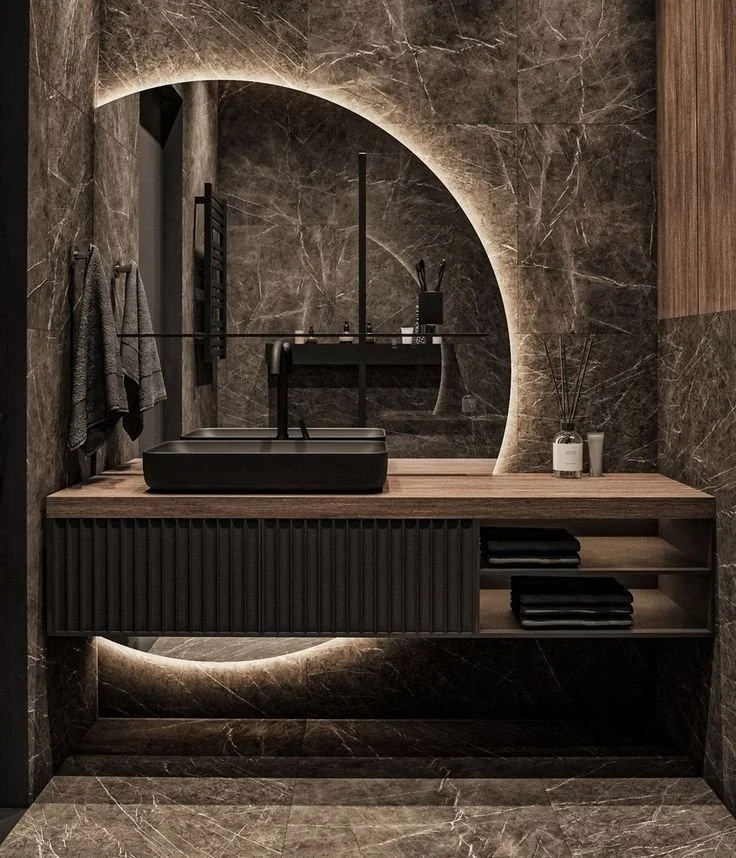 A modern bathroom vanity with a black vessel sink, black faucet, and a large circular backlit mirror. The wall and floor are covered in dark marble, and there are towels and bathroom essentials on the wooden countertop and open shelf.