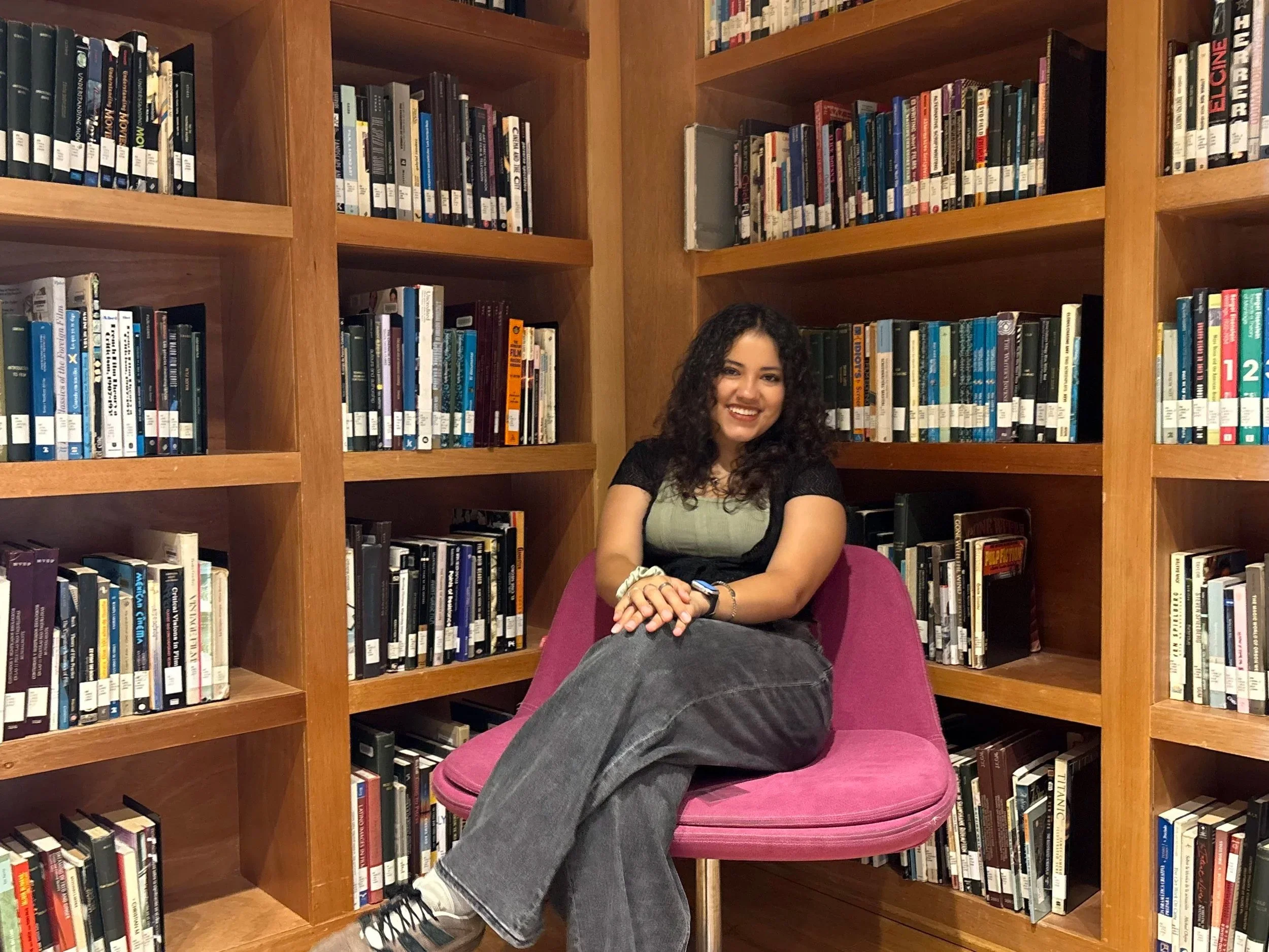 A young woman with curly dark hair smiling and sitting with her legs crossed on a pink chair in a library. She is wearing a light-colored top and gray pants, with a smartwatch on her wrist. The background consists of wooden bookshelves filled with a variety of books.