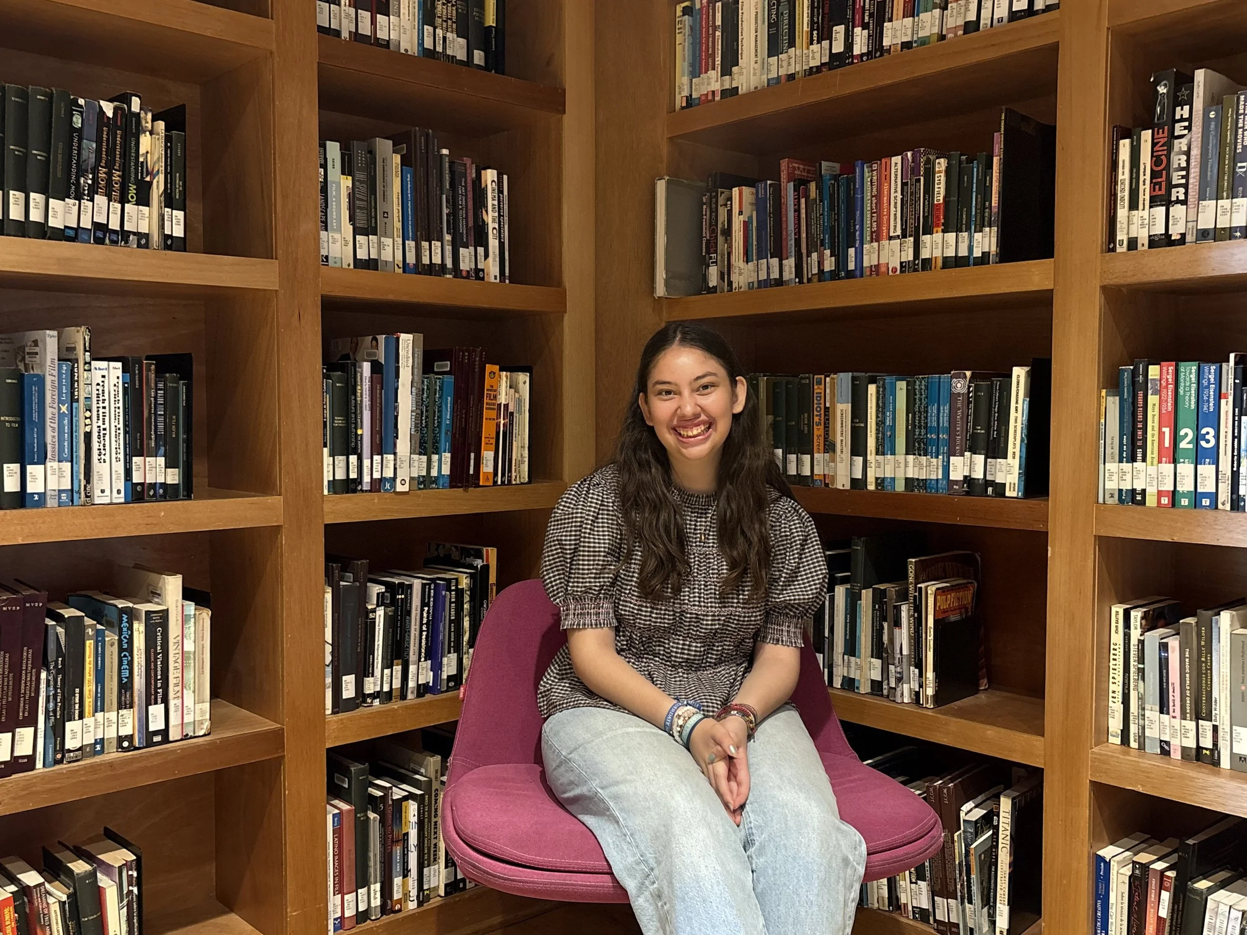 A young woman with long dark hair, smiling, sitting on a pink chair in front of wooden bookshelves filled with books in a library.