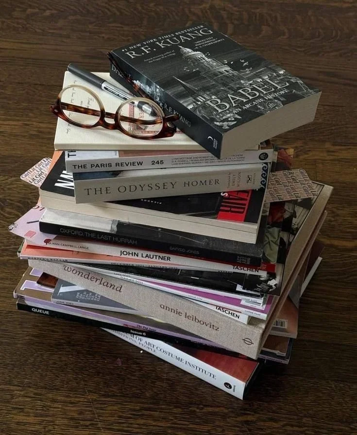 A stack of books on a wooden surface, with reading glasses resting on top. The top book is "Babylon" by R.F. Kuang.